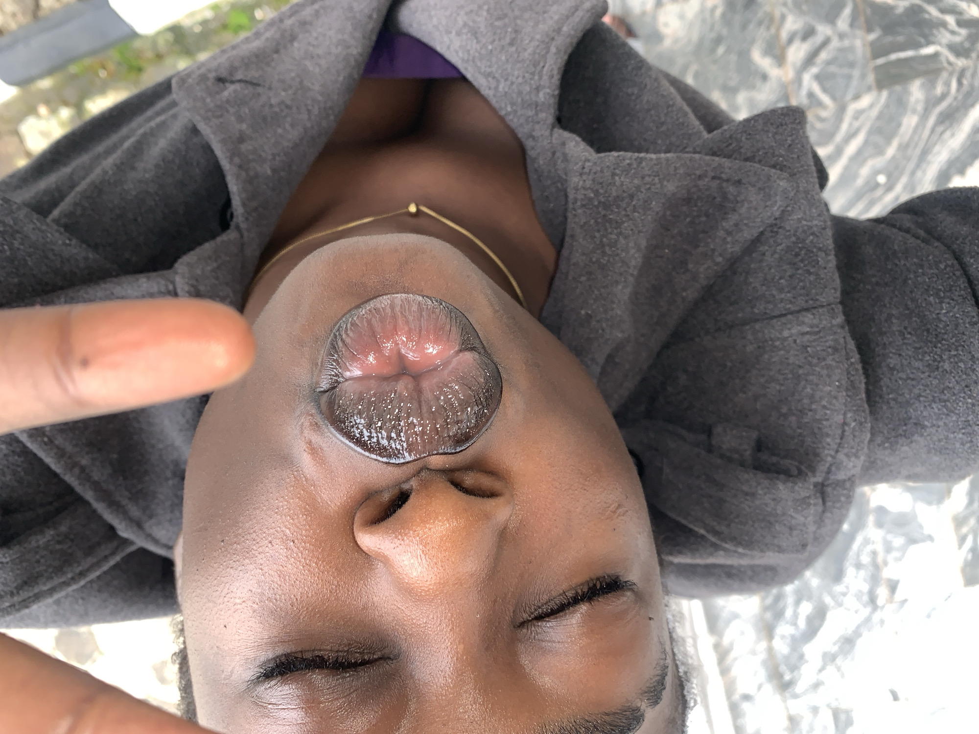 The image is a close-up, low-angle, overhead shot of a person's face from the nose down, with their eyes closed and lips parted. The person has dark skin and is wearing a grey hooded garment that covers their neck and chest. A thin gold chain with a small bead is visible around their neck. Their lips are wet and appear to have a glossy or oily sheen. The background is blurred but appears to be an outdoor setting with a patterned surface that could be pavement or tiles, and some greenery is visible. The image is taken in Gura Top, Nigeria.