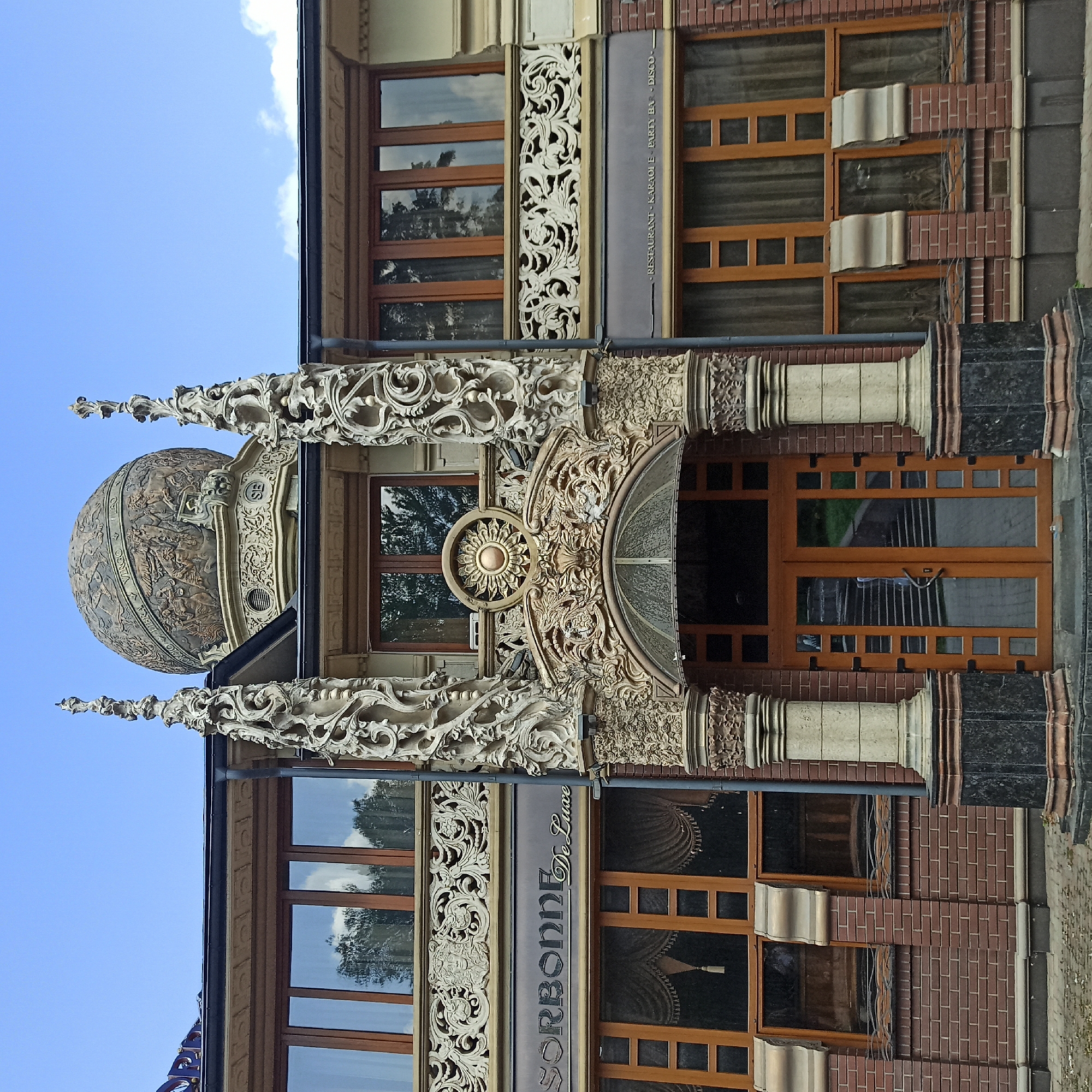 The image shows the facade of a building with ornate architectural details. The central entrance is framed by elaborate carved columns, a decorative archway with a sun-like motif, and a glass awning. Above the entrance, a large, spherical dome is visible, decorated with bas-reliefs depicting zodiac signs and mythological figures. The building has multiple windows with wooden frames, some with draped curtains. The facade is constructed with red brick and features decorative friezes and cornices. A sign above the entrance reads "SORBONNE De Luxe". Another smaller sign below the upper windows reads "RESTAURANT KARAOKE PARTY BAR DISCO". The building is located on a cobblestone street. The sky above is blue with scattered clouds. This building is known as the Sorbonne Restaurant and is located in Chernivtsi, Ukraine. There are no people visible in the image.