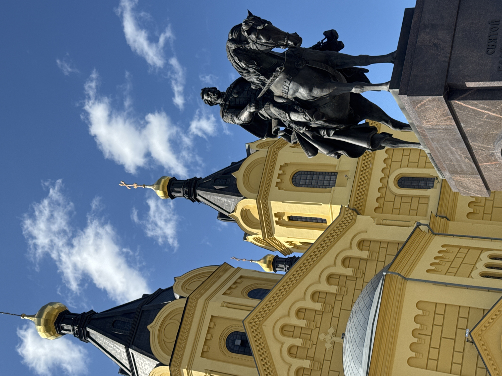 The image shows a bronze equestrian statue in front of the Alexander Nevsky Cathedral in Nizhny Novgorod, Russia. The cathedral is a prominent yellow building with gold domes and black spires, characteristic of Russian Orthodox architecture. The sky is a clear blue with scattered white clouds. The statue depicts a man in historical armor on horseback, facing to the right. The statue is placed on a dark, multi-tiered granite pedestal. The foreground shows the base of the pedestal and the statue's legs, while the background features the impressive facade of the cathedral with its distinct domes and windows. The lighting suggests a sunny day.
