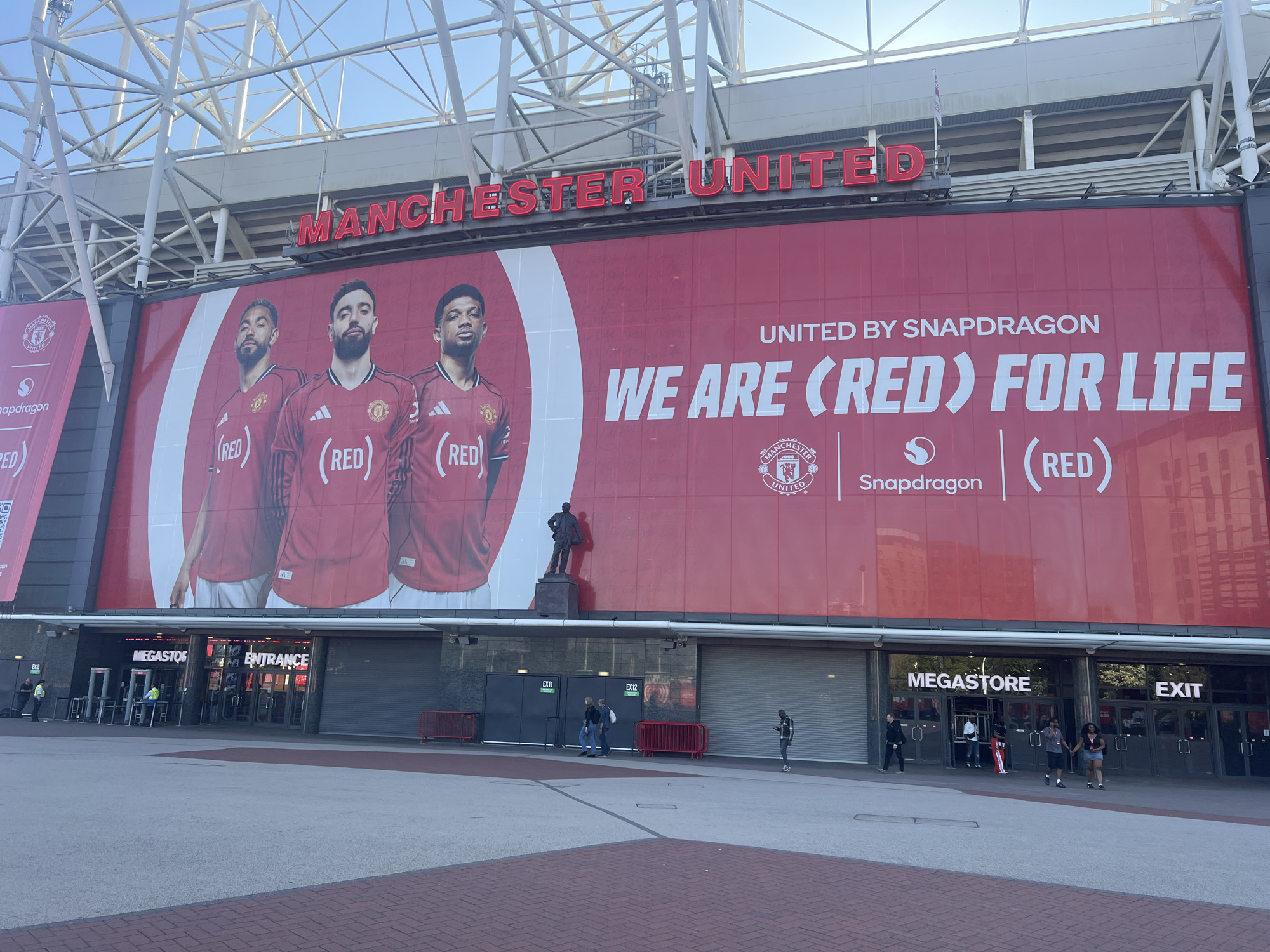 Here is a detailed description of the image:
The image shows the exterior of Old Trafford stadium, home to Manchester United, located in the city of Trafford, United Kingdom. A large, red banner spanning the length of the stadium is visible above the ground-level entrances. The banner features the Manchester United logo, three soccer players in red uniforms, and the text "UNITED BY SNAPDRAGON," "WE ARE (RED) FOR LIFE," and "(RED)" alongside the Snapdragon logo. A statue stands in front of the banner.
Below the banner are several storefronts, including the "MEGASTORE" and "ENTRANCE" to the stadium. A few people are seen walking around and entering/exiting the building. The pavement in front of the stadium is made of grey concrete with a red brick border. Above the banner, the stadium's structural supports are visible against the sky.