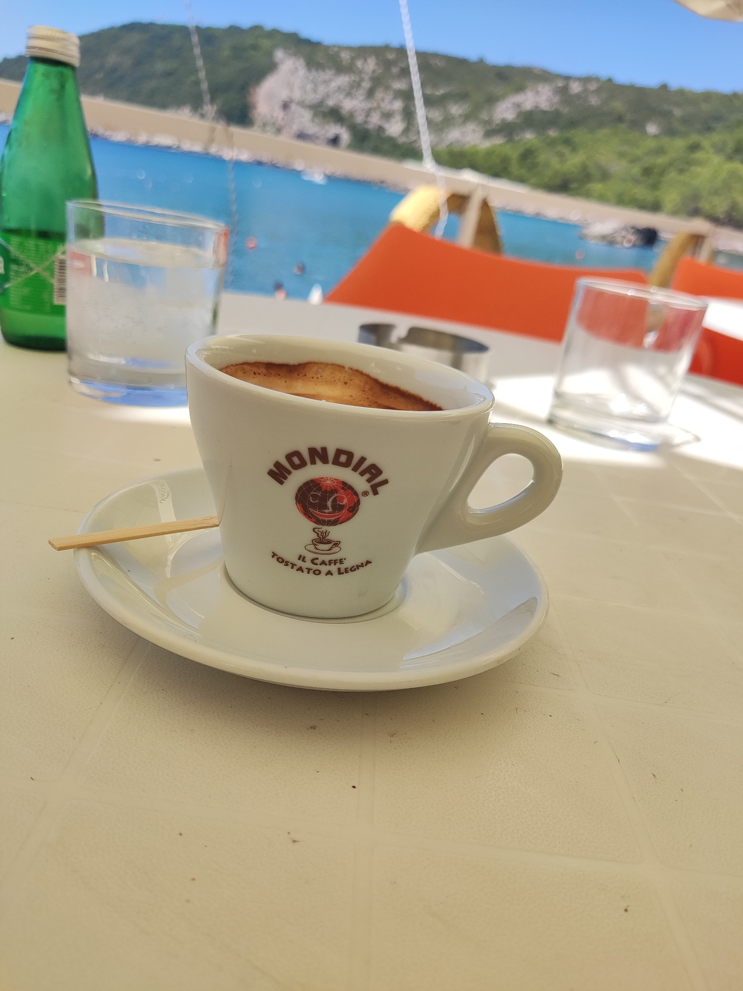 A close-up, eye-level shot captures a white ceramic coffee cup filled with dark espresso, placed on a saucer. A small wooden stirrer rests across the saucer and the rim of the cup. The cup bears a red and black logo with the text "MONDIAL" and "IL CAFFÈ TOSTATO A LEGNA". In the background, out of focus, is a body of water with several small boats and swimmers, leading to a tree-covered hill and a clear blue sky. To the left of the coffee cup are two glasses, one filled with water and ice, and a green bottle. Orange seating is visible further in the background. The scene appears to be an outdoor cafe or restaurant setting, likely in Zagrađe, Montenegro, given the visual elements suggestive of a coastal Mediterranean location. The table surface is a textured white cloth.