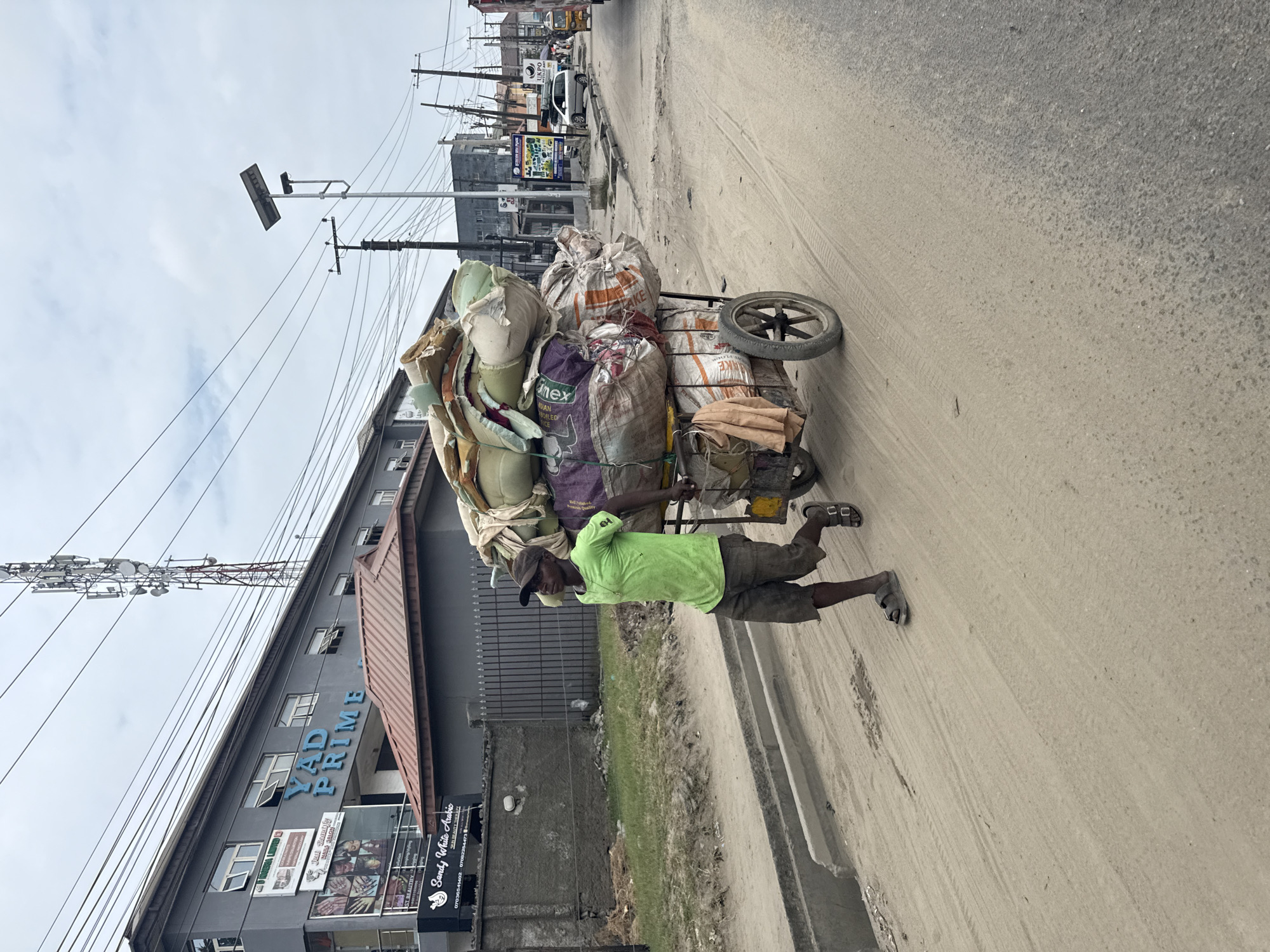 A man in a green t-shirt and dark shorts is pushing a two-wheeled cart loaded with various sacks and what appears to be foam padding. He is walking on a dusty, unpaved road. In the background, there is a grey building with "YAD PRIME" written on it in blue letters, and beyond that, a cell tower. Further in the distance, there are more buildings, utility poles, power lines, and a few vehicles on a paved road. The scene suggests an urban environment in Nigeria, likely Ado, characterized by a mix of commercial buildings, infrastructure, and informal transport. The sky is overcast.