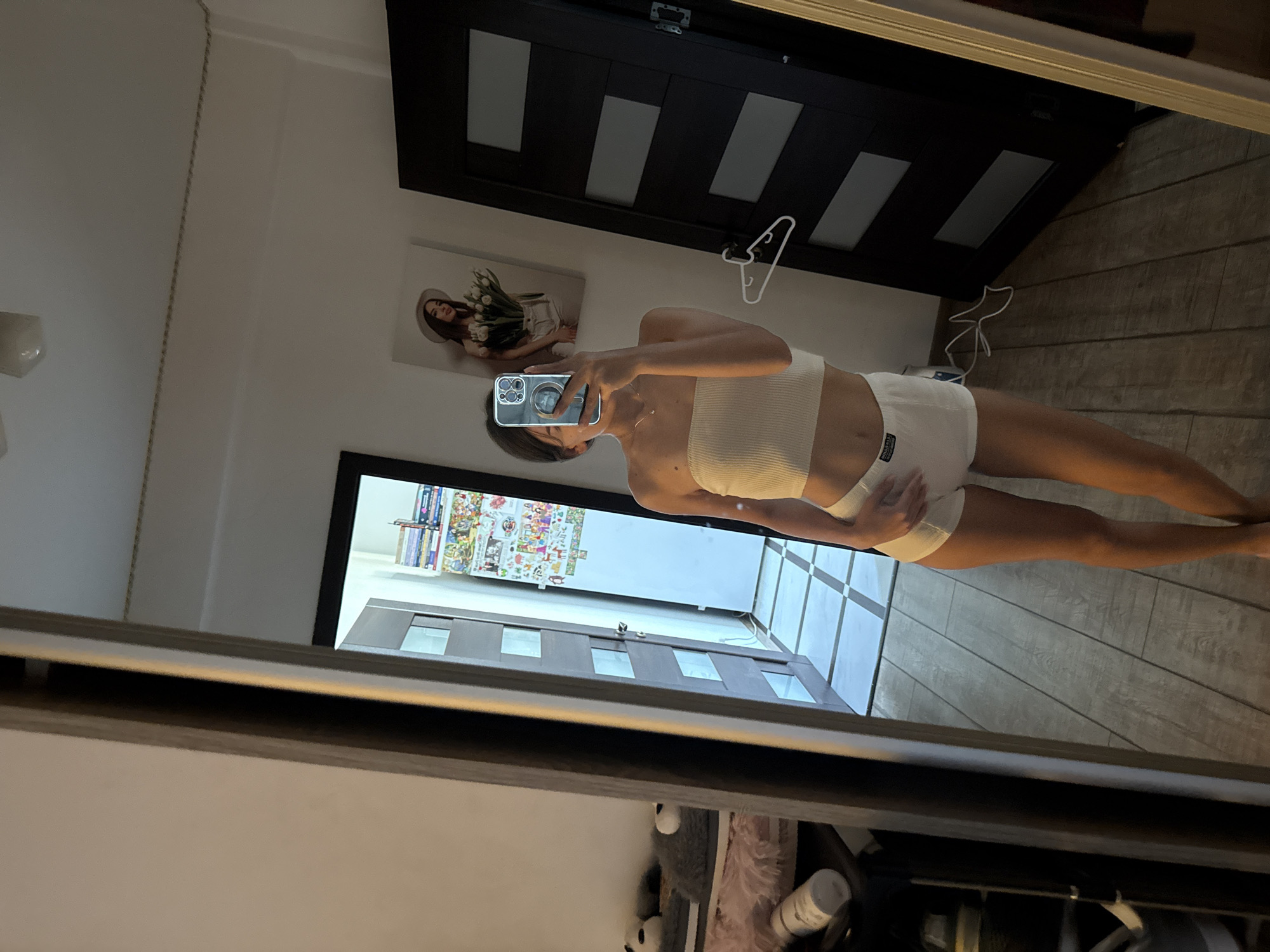 The image shows a young woman taking a selfie in a mirror. She is wearing a white ribbed bandeau top and white shorts. The setting appears to be a bedroom, with a door and a picture on the wall visible in the reflection. The floor is made of wooden planks. The overall impression is that of a casual, indoor setting. There is no indication of the location being in Chișinău, Moldova.