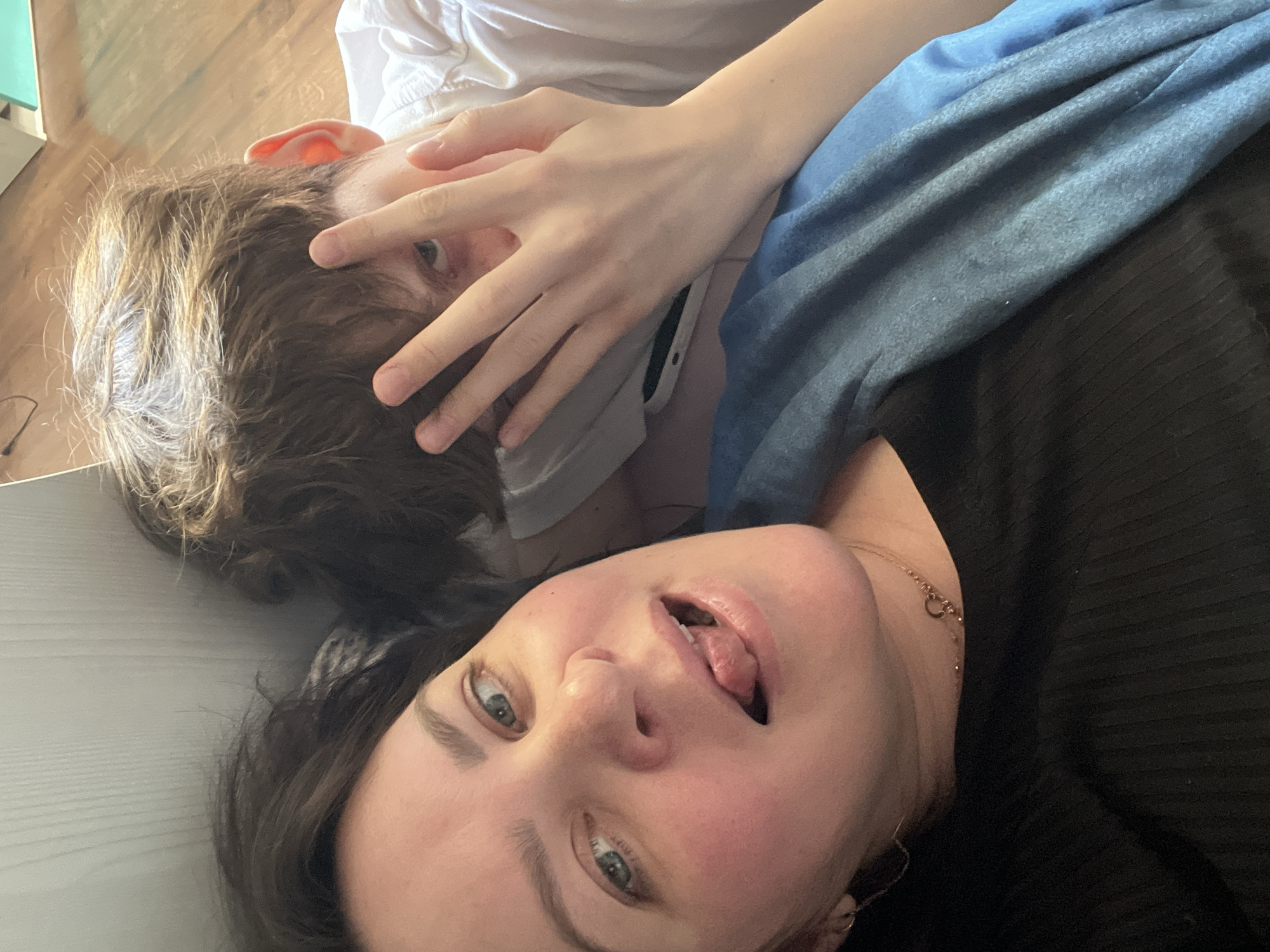 The image shows two people lying down indoors. One person, likely female, is close to the camera, with dark hair and green eyes. She has her mouth open, sticking her tongue out. She is wearing a black ribbed shirt and a gold necklace. The other person has brown hair, is partially obscured by a hand and a white face mask, and is wearing a white t-shirt. A blue denim-like fabric covers part of them. They appear to be lying on a light-colored floor or surface. The location of Noviy, Russia is not discernible from this image.
