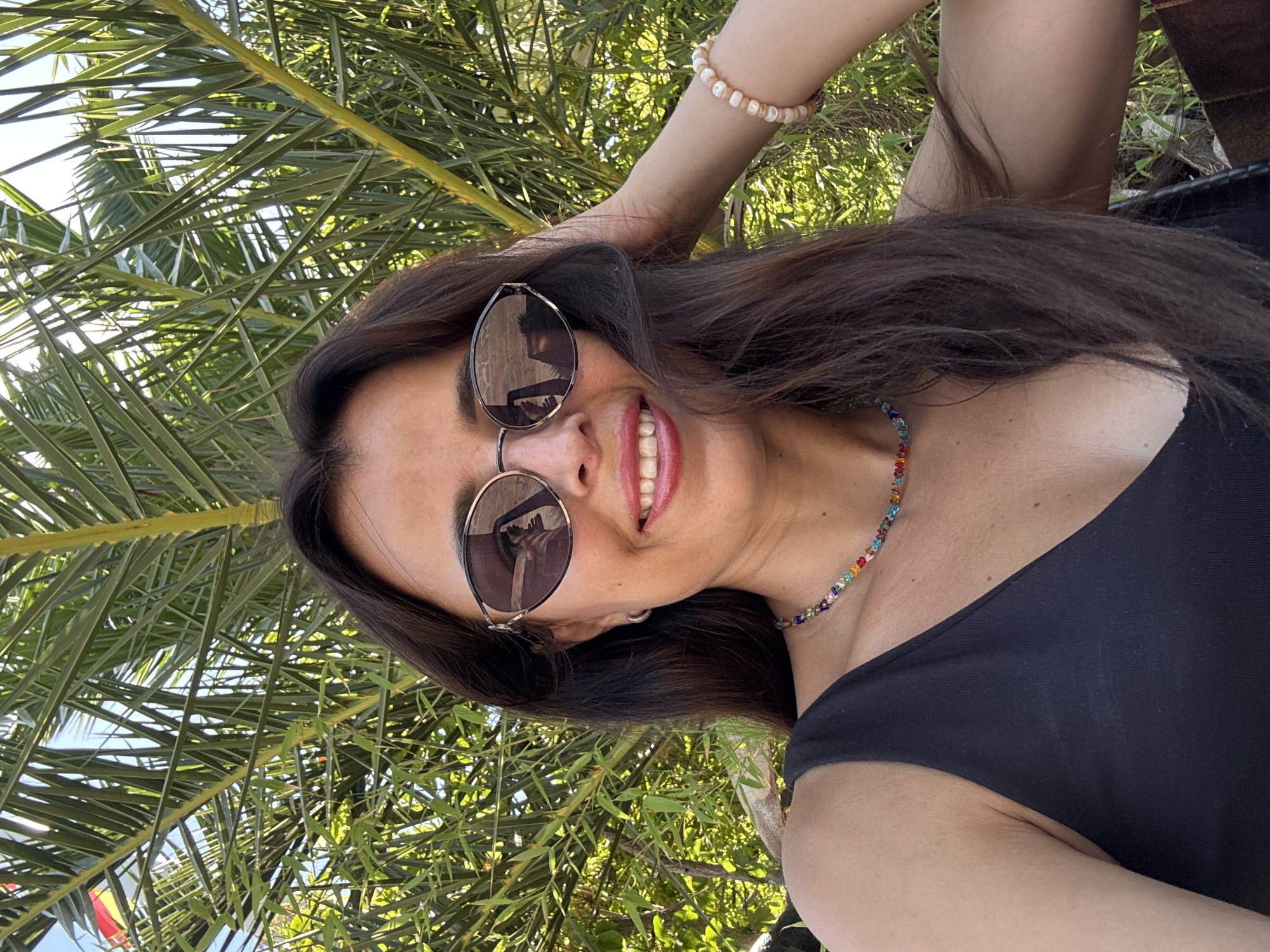 A woman is shown in a close-up selfie, with her head tilted and her hair falling around her shoulders. She is wearing a black, one-shoulder top and a colorful beaded necklace. Her sunglasses are perched on her nose, reflecting the scene around her. She has a bright smile, with her teeth showing, and her left arm is raised and resting on her head. She is wearing a pearl bracelet on that arm. The background is filled with lush green palm fronds, suggesting an outdoor, tropical setting. Sunlight filters through the leaves, casting dappled shadows and creating a warm, bright atmosphere. The image appears to be taken in daylight, likely in the late morning or afternoon. The woman's relaxed posture and joyful expression suggest a feeling of happiness and contentment.