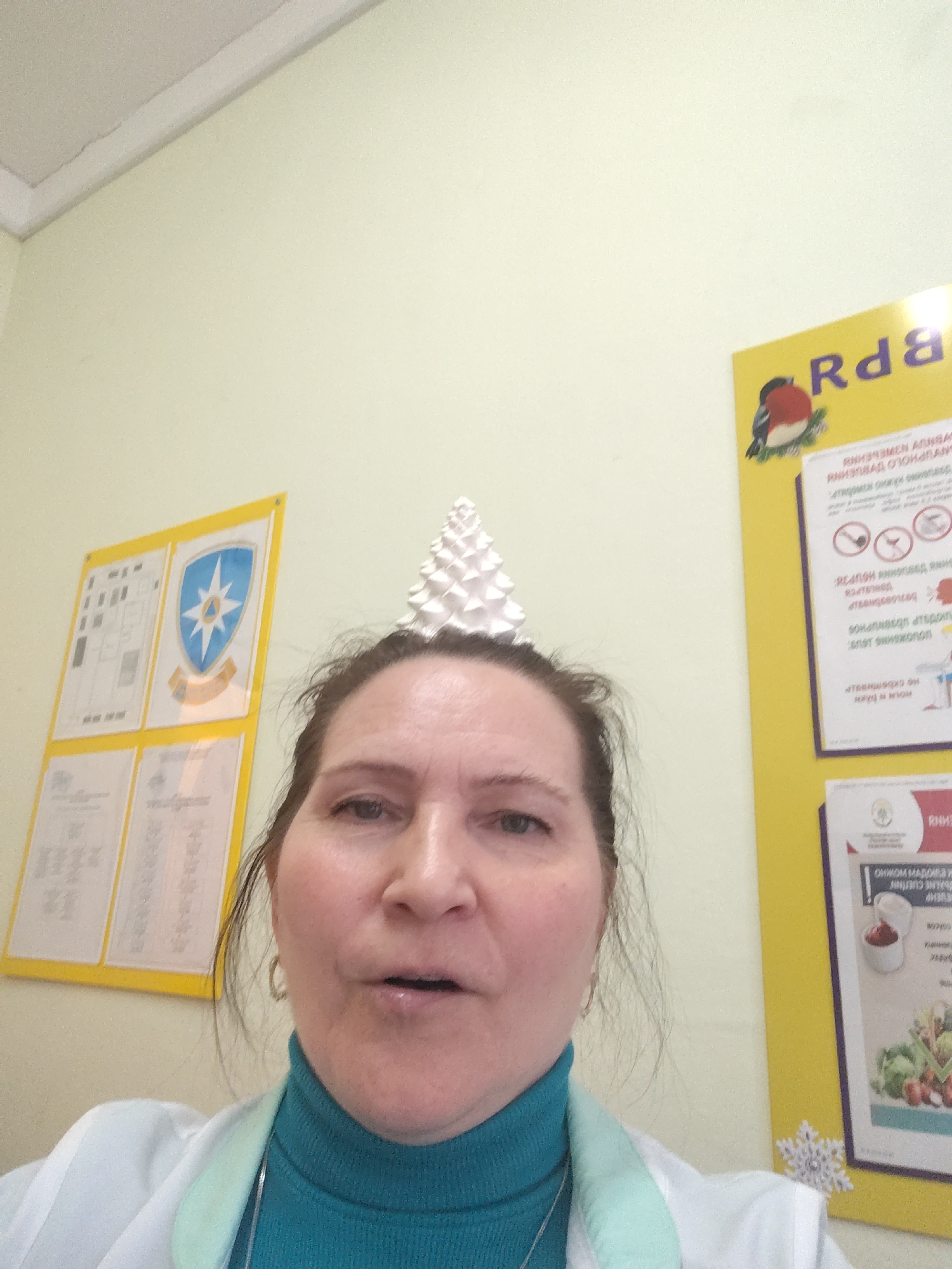A woman in a turquoise turtleneck and white lab coat stands indoors, likely in a medical or educational setting, given the informational posters on the yellow walls behind her. A decorative, white cone-shaped object sits atop her head, perhaps as a playful headdress. One poster features a coat of arms or emblem, while another has text that appears to be Cyrillic. The environment suggests a public facility in Sarapul, Russia.