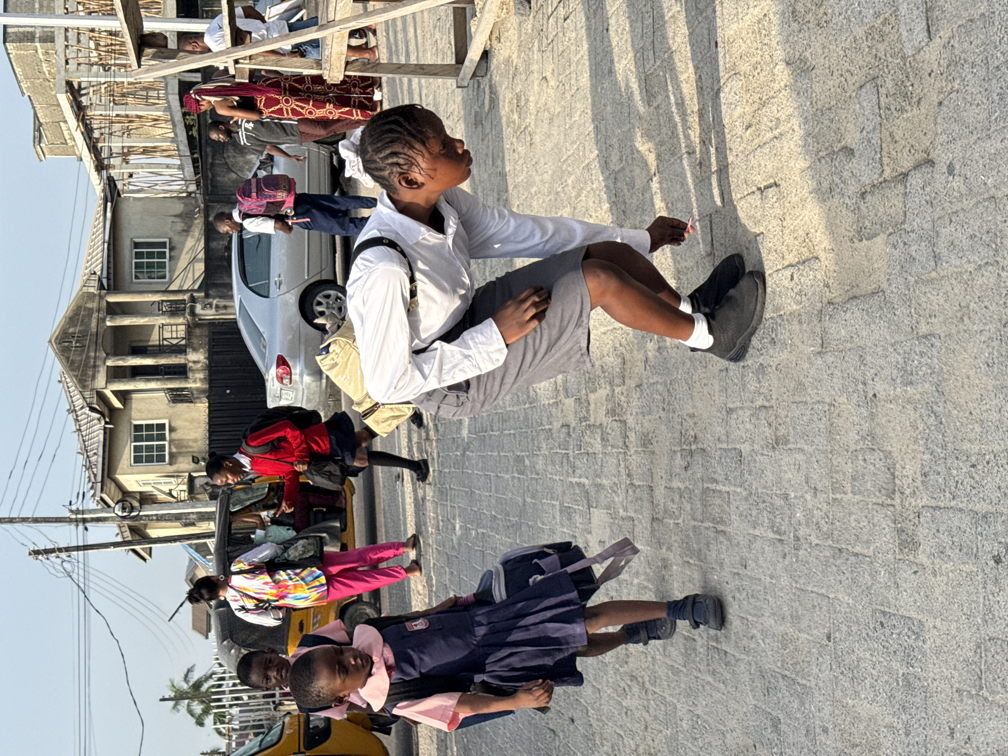 A slightly high-angle, full daylight shot shows an African street scene with multiple people in the city of Ado, Nigeria. In the midground, a dark-skinned young woman is crouched with her legs extended and holding a small object in her right hand. She wears a white, button-down shirt, a gray skirt, and black shoes with white socks. A backpack is slung over her left shoulder.
Behind and to the left of the woman, other young people are walking along the street, some wearing school uniforms. A yellow auto rickshaw is visible. Behind the rickshaw, more young people are walking, some with backpacks. A light gray car is parked nearby, with other people walking behind it.
In the background, a building under construction is visible along with additional buildings. The street is paved with interlocking, gray stone blocks, casting long shadows in the strong sunlight. Power lines and a palm tree are partially visible in the distance.