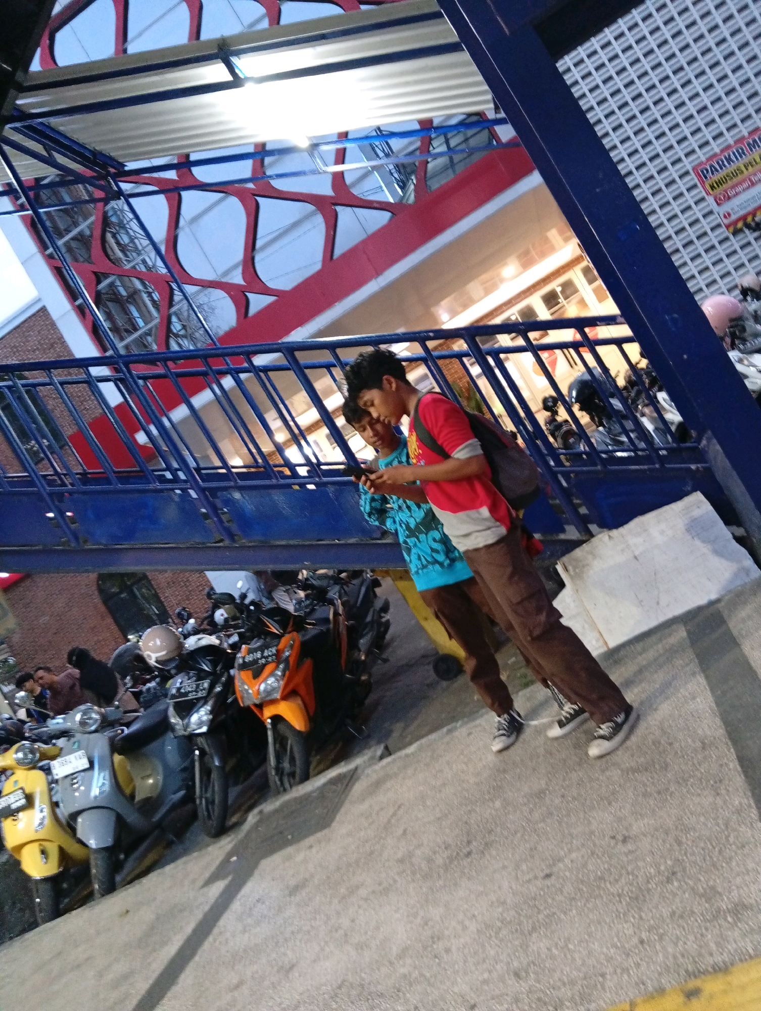 The image captures a scene in Malang, Indonesia, depicting a pedestrian overpass with two young men in the foreground. One man, wearing a red and gray t-shirt and brown pants, is looking at his phone. The other, in a blue t-shirt with white lettering and brown pants, is also looking at his phone. Both are wearing black and white sneakers. Behind them, a blue metal railing separates them from a row of parked motorcycles, including a yellow scooter, a gray scooter, a black motorcycle, and an orange motorcycle. The overpass structure is visible above, with its red and white diagonal supports and a corrugated metal roof. In the background, a wall with a grid pattern and a sign that reads "PARKIR MOTOR KHUSUS PELAJAR" (Motorcycle Parking for Students) is partially visible, suggesting the location is near an educational institution. The ground is paved with concrete, and shadows are cast by the overpass and the motorcycles. The overall setting suggests a public area within the city.