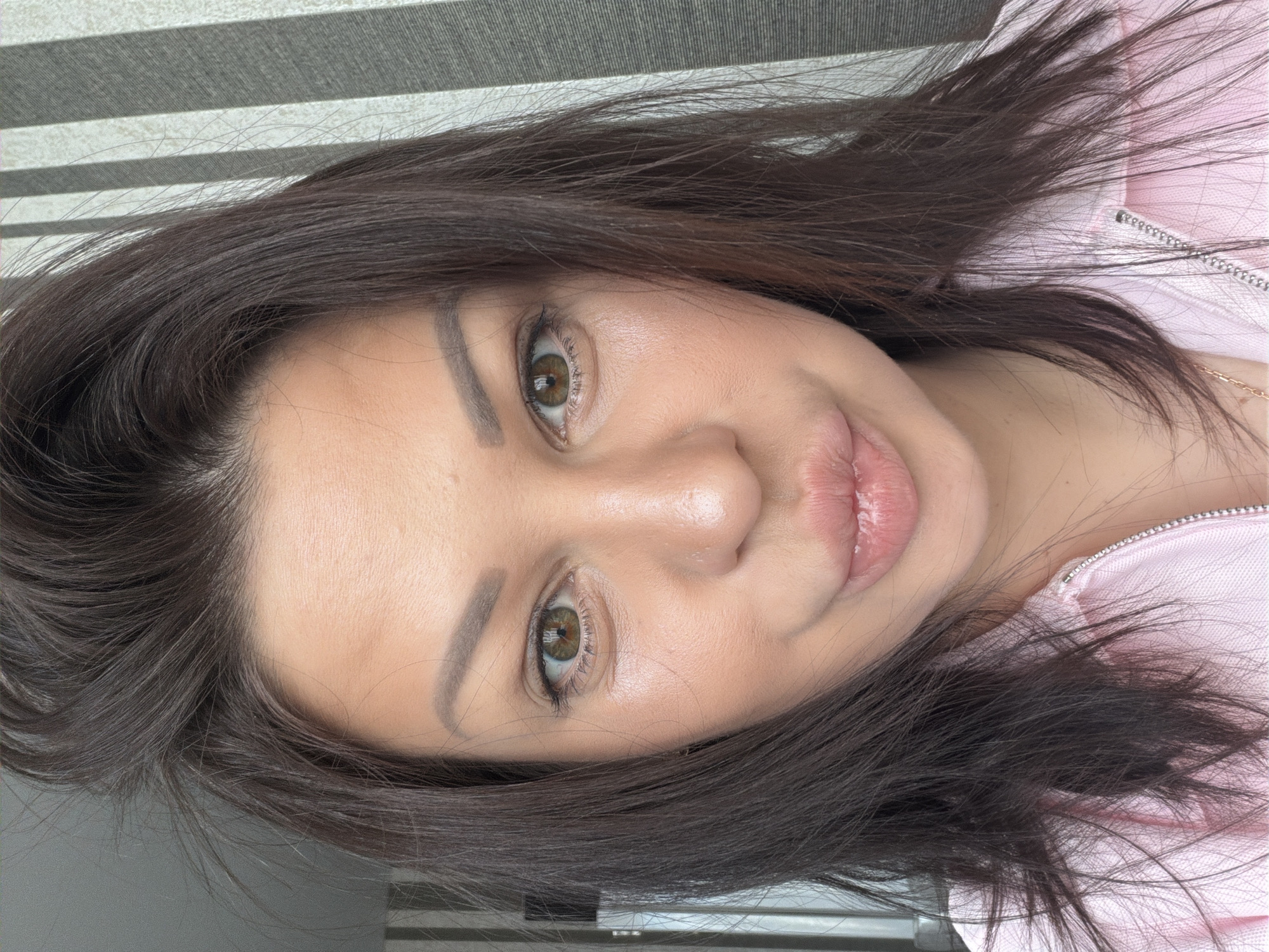 A close-up selfie of a woman with brown hair and brown eyes, looking at the camera. She is wearing a pink zippered jacket. Her hair is messy and falls around her face. She is puckering her lips. The background consists of vertical striped curtains. The image appears to be taken indoors, possibly in an apartment or hotel room. There is no visible indication of the city of Sochi, Russia.