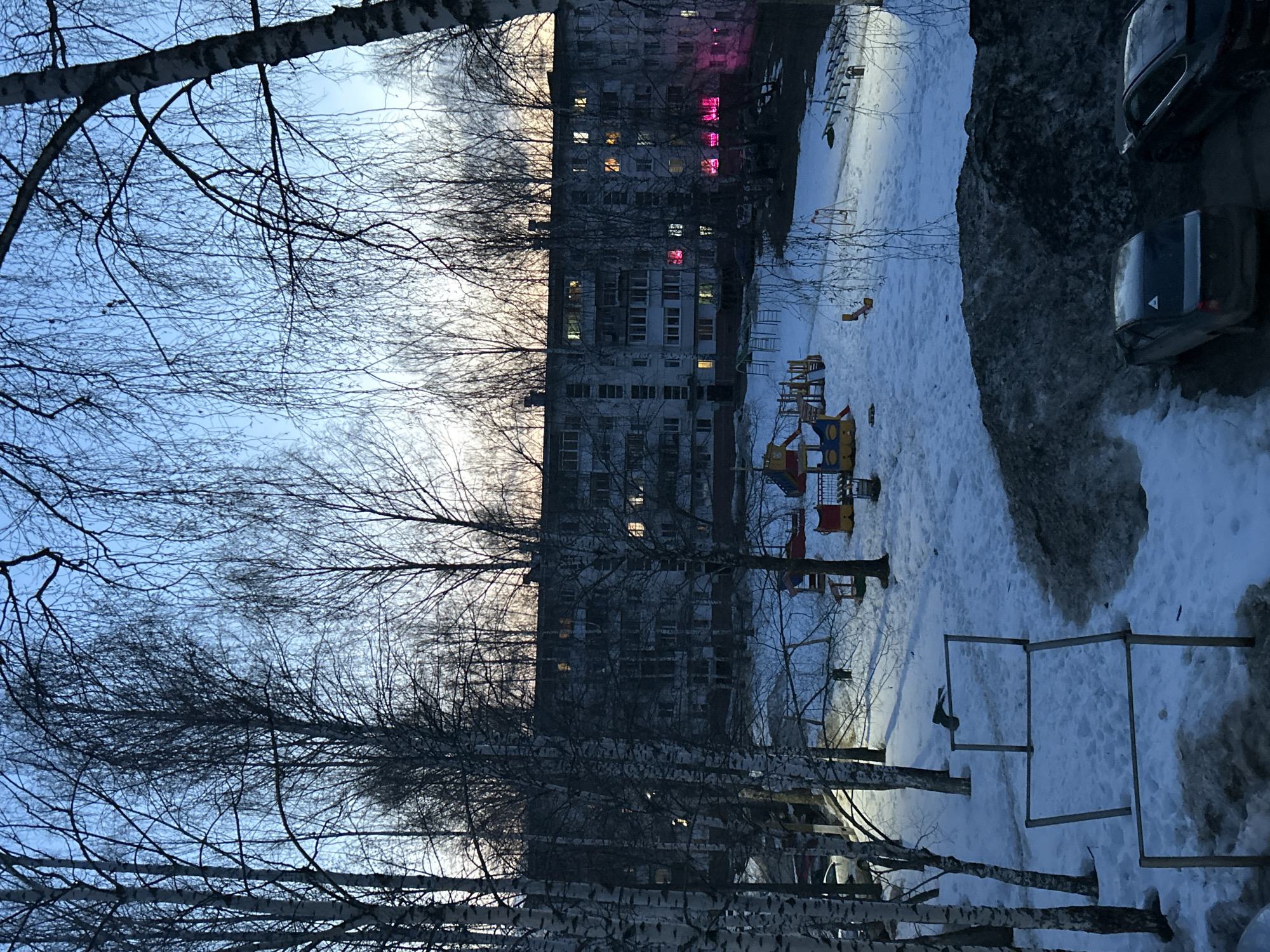 The image shows a snowy courtyard in Chaikovsky, Russia, at dusk. A multi-story apartment building is in the background with some illuminated windows, including some with pink lights. In the foreground are bare birch trees, a snow-covered playground, and a metal structure. Two cars are visible on the right. There are no people visible in the image. The environment appears to be a residential area with typical Soviet-era apartment buildings.