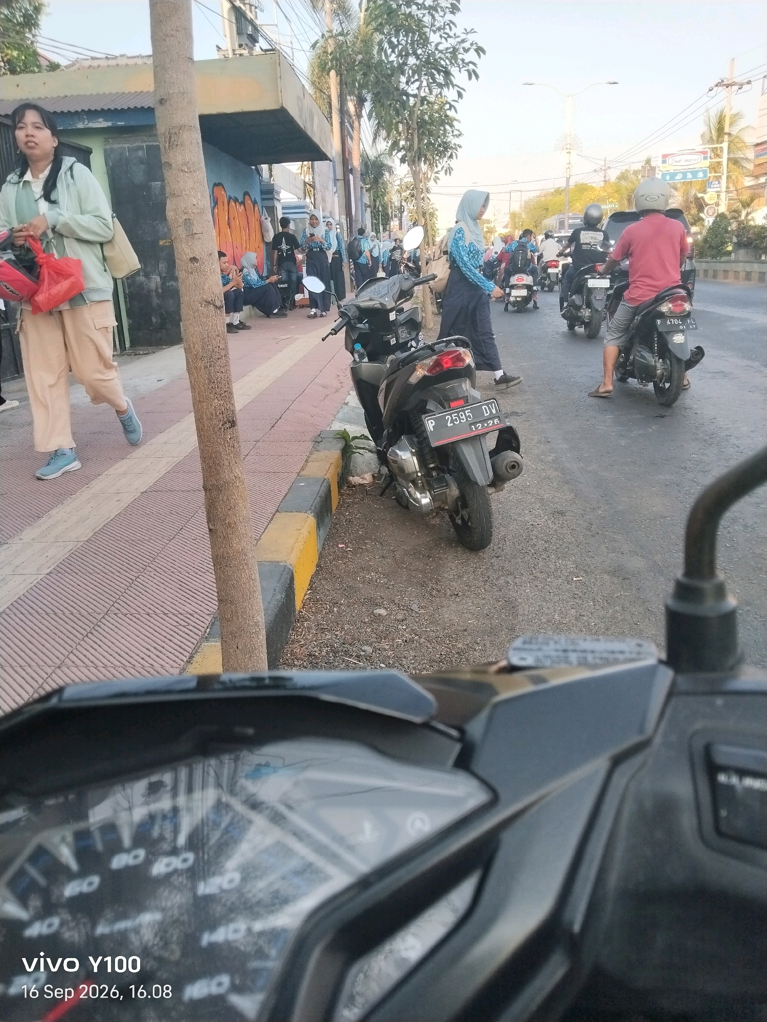 The image captures a street scene in Situbondo, Indonesia, with a focus on a parked motorcycle in the foreground and other motorcycles and pedestrians on the street and sidewalk. The perspective is from a motorcycle rider, as indicated by the close-up view of a motorcycle's dashboard and handlebars.
In the mid-ground, several people are visible. On the sidewalk to the left, a woman wearing a light green jacket, beige pants, and blue sneakers walks while carrying red bags and a beige purse. Further down the sidewalk, a group of young people, likely students, are seated or standing. Some are wearing blue and white uniforms, with a few wearing headscarves.
On the street, multiple motorcycles are present. One motorcycle, with license plate P 2595 DV, is parked on the side of the road, facing away from the camera. Another motorcycle, with license plate P 4704 FL, is also visible further down the street with a rider wearing a red shirt and a helmet. Traffic is moving on the road, with several other motorcycles visible in the distance.
The environment includes a paved sidewalk with a decorative pattern, a curb painted with yellow and black stripes, and a dirt and gravel shoulder. A tree trunk stands prominently on the left side of the frame, partially obscuring the sidewalk and a building in the background. The building has a light-colored facade and a dark-colored roof. Sunlight suggests it is daytime, likely late afternoon given the timestamp on the image. The sky is clear. The general atmosphere appears to be one of everyday urban life, with people commuting and gathering.