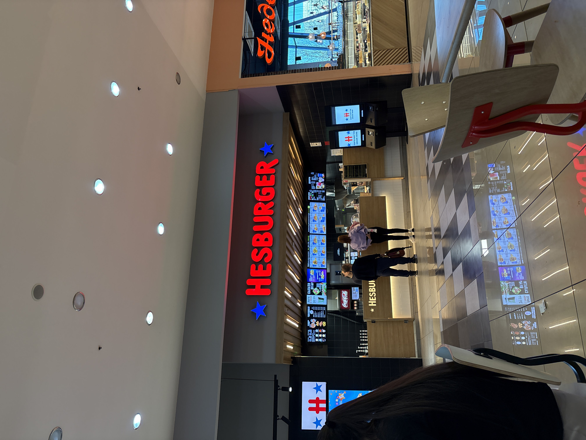 The image shows the interior of a shopping mall, specifically focusing on the "Hesburger" fast-food restaurant. The Hesburger logo is prominently displayed in red neon letters with a blue star on either side. There are menu boards showing various food items and prices. A few customers are visible near the counter. In the upper portion of the image, there is another restaurant. The setting appears to be a modern shopping mall with a tiled floor. The location of the city is Pleven, Bulgaria.