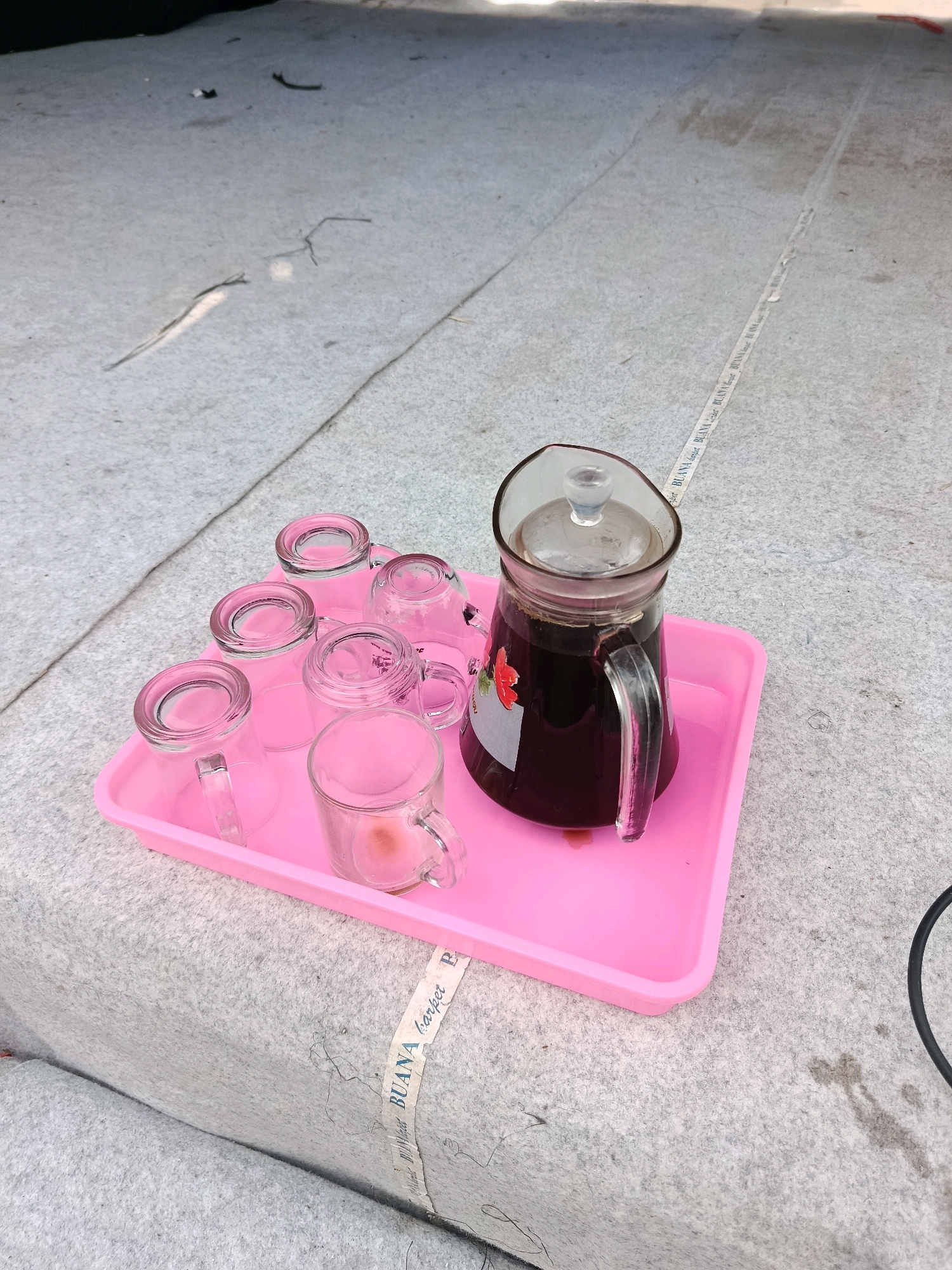 A pink plastic tray holds a glass pitcher filled with dark liquid, possibly coffee, and six small glass cups. The tray and its contents are situated on a light-colored, textured surface, possibly a carpet or mat, with a strip of white tape bearing blue text, including "BUANA carpet," visible on it. The scene is outdoors, with dappled sunlight creating shadows. The location is identified as Bululawang, Indonesia. There are no people or animals visible in the image.