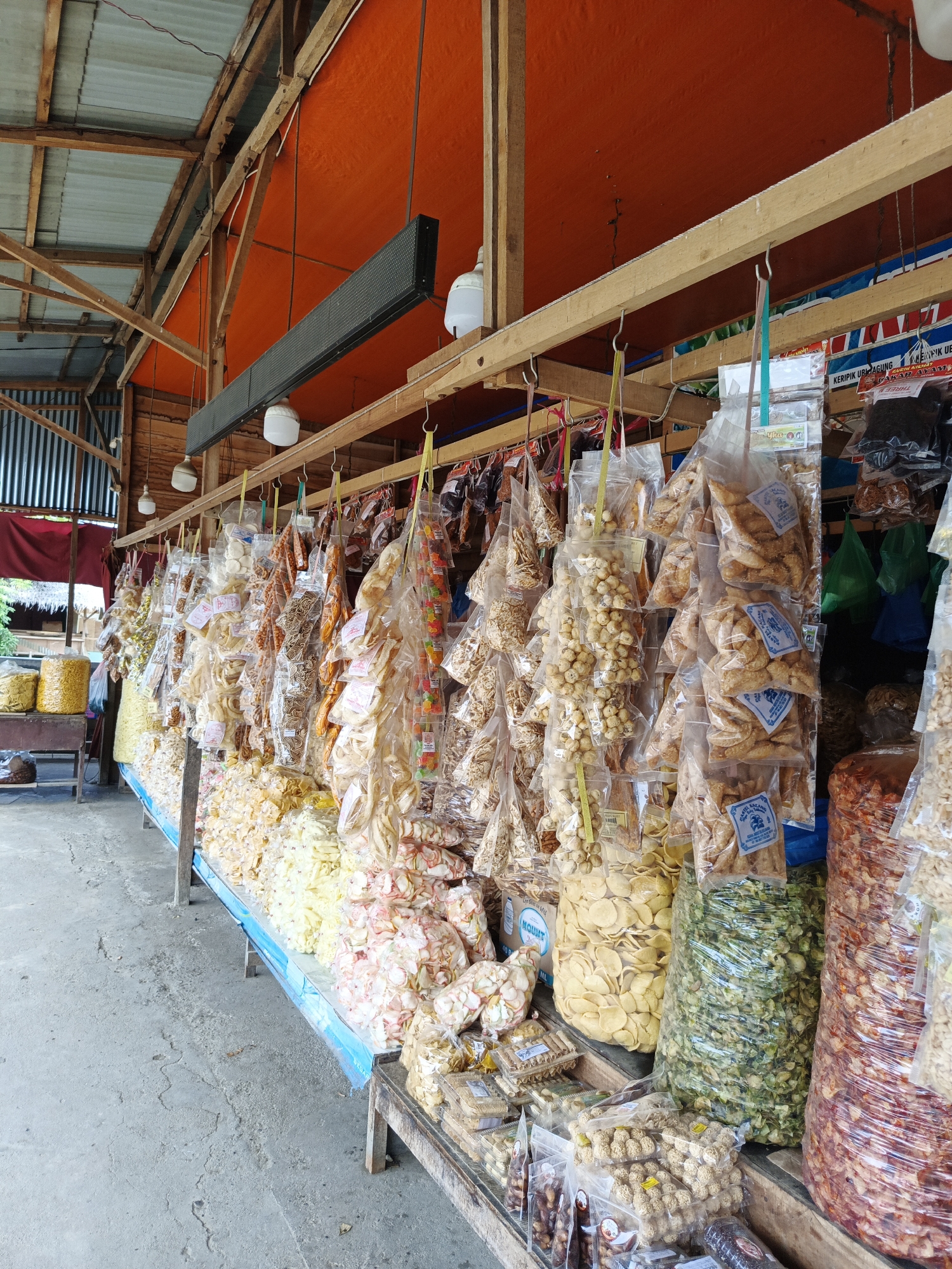 The image depicts a stall selling a wide variety of snacks, likely in Sawang, Indonesia. The stall is set up outdoors, under a wooden structure with an orange tarp for a roof, suggesting a market or roadside vendor. Various snacks are displayed, mostly in clear plastic bags, hanging from hooks or arranged on shelves and tables. These include an assortment of chips, crackers, nuts, and what appear to be puffed rice snacks, with colors ranging from pale yellow and white to green and reddish-brown.
Some of the snacks are packaged in large, clear plastic sacks, while others are in smaller, individually sealed bags. Several bags have labels with text, though most are too small or blurry to be read clearly. One prominent label, visible on several bags of triangular-shaped snacks, features a blue design with white text. Large bags of light yellow, green, and reddish-brown snacks are particularly noticeable due to their size.
In the background, there are glimpses of corrugated metal siding and some greenery, hinting at the surrounding environment. A long, black digital display board is mounted above the stall. The lighting suggests it is daytime, possibly late morning or afternoon, with diffused sunlight. The overall impression is that of a bustling market scene with a vibrant display of local food products. There are no people visible in the immediate foreground or actively engaged in transactions, focusing the view solely on the merchandise.