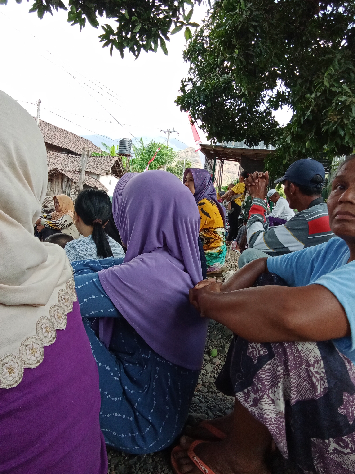In Trebungan, Indonesia, a group of people are gathered outdoors, seated on the ground. In the foreground, a woman in a light-colored hijab and a purple garment is on the left, her embroidered trim visible. To her right, another woman in a purple hijab and a blue patterned skirt is seated. In the midground, a man wearing a blue cap and a striped shirt is visible, his hands clasped. Behind him, other individuals are seated, including a woman in a yellow and green patterned hijab. The environment suggests a rural or semi-rural setting with a tiled-roof building and trees in the background. The ground appears to be covered with small stones and dirt. The overall impression is of a community gathering.
