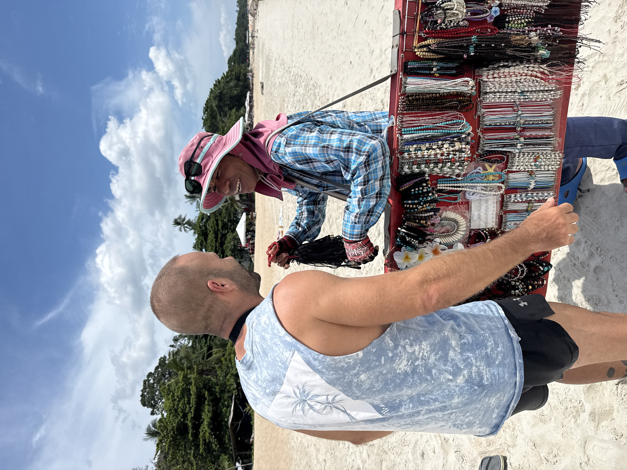 A man with a shaved head, wearing a light blue sleeveless t-shirt with a palm tree design and black athletic shorts, stands on a sandy beach in the Ko Samui District of Thailand. He is facing a vendor, a person wearing a pink bucket hat and sunglasses, a blue and pink plaid shirt, and a pink neck covering. The vendor is smiling and holding a bundle of black beaded necklaces. The vendor's merchandise, a display of colorful beaded necklaces and bracelets, is arranged on a red cart between them. The beach stretches out behind them, with palm trees and vegetation in the background. The sky is blue with scattered white clouds.