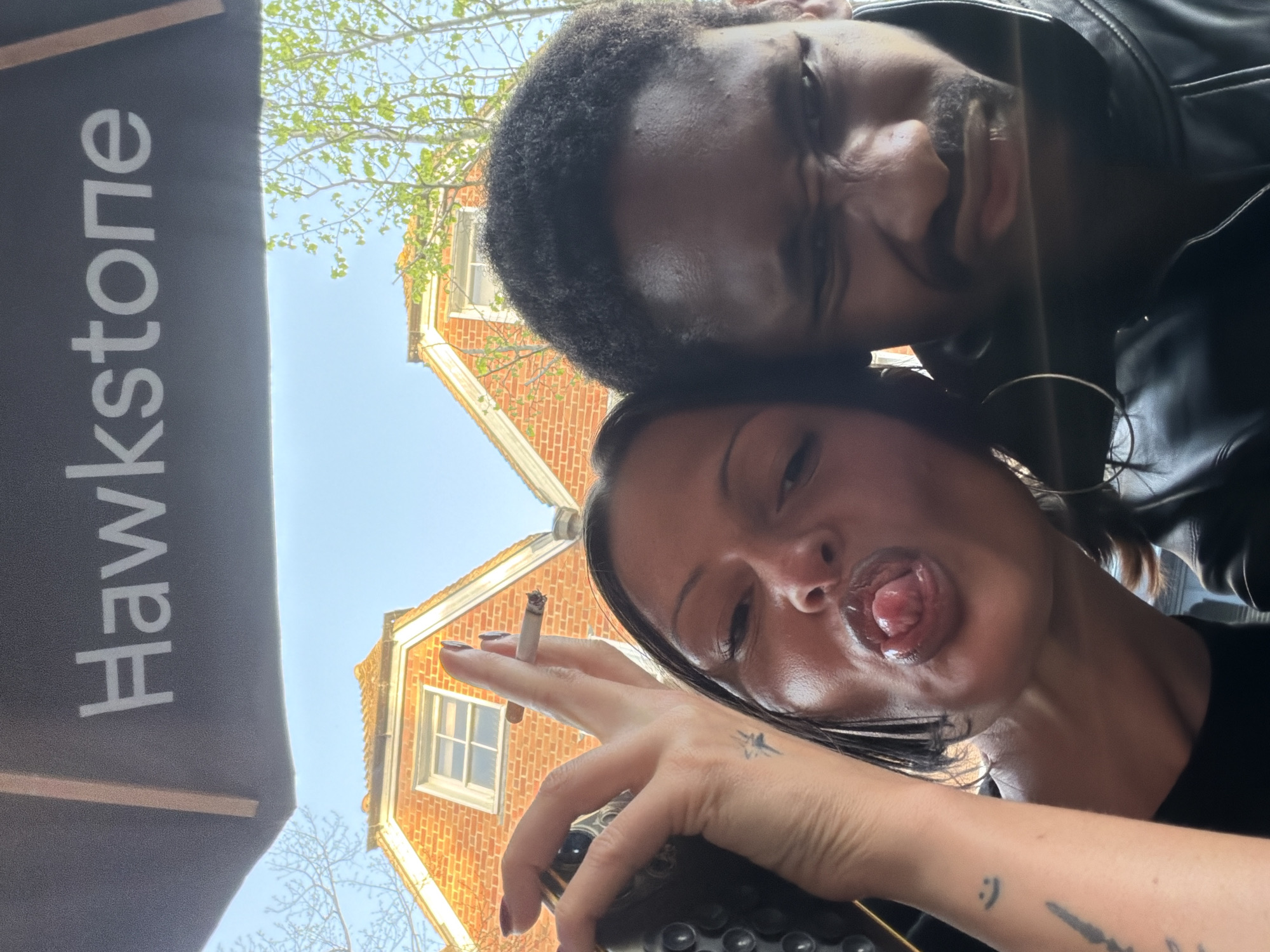 Two individuals, a man and a woman, are positioned close together, taking a selfie. The woman has her tongue extended and is holding a lit cigarette. The location is identifiable as Tunbridge Wells, United Kingdom, due to the presence of a "Hawkstone" sign, a brand of cider linked to Tunbridge Wells. The background includes a building with red brickwork and a window. The sky is visible above the building.