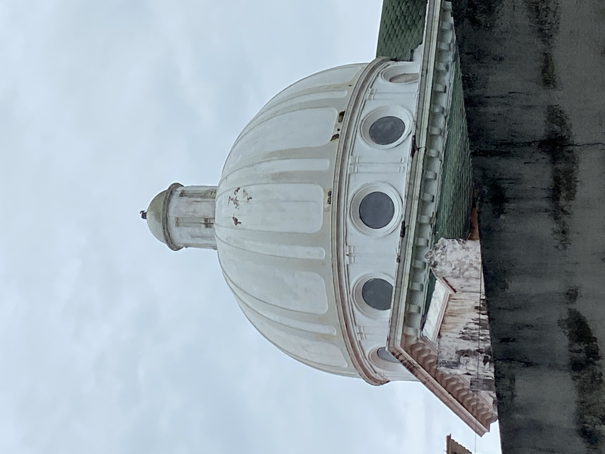 An upward-angled view shows a building dome with circular windows against a cloudy sky. The dome is painted white and has vertical ridges. Three evenly-spaced circular windows line the midsection of the dome. The dome is attached to a lower structure with a green tiled roof. A concrete wall runs vertically on the right side of the frame. The location is Río Chico, Venezuela.