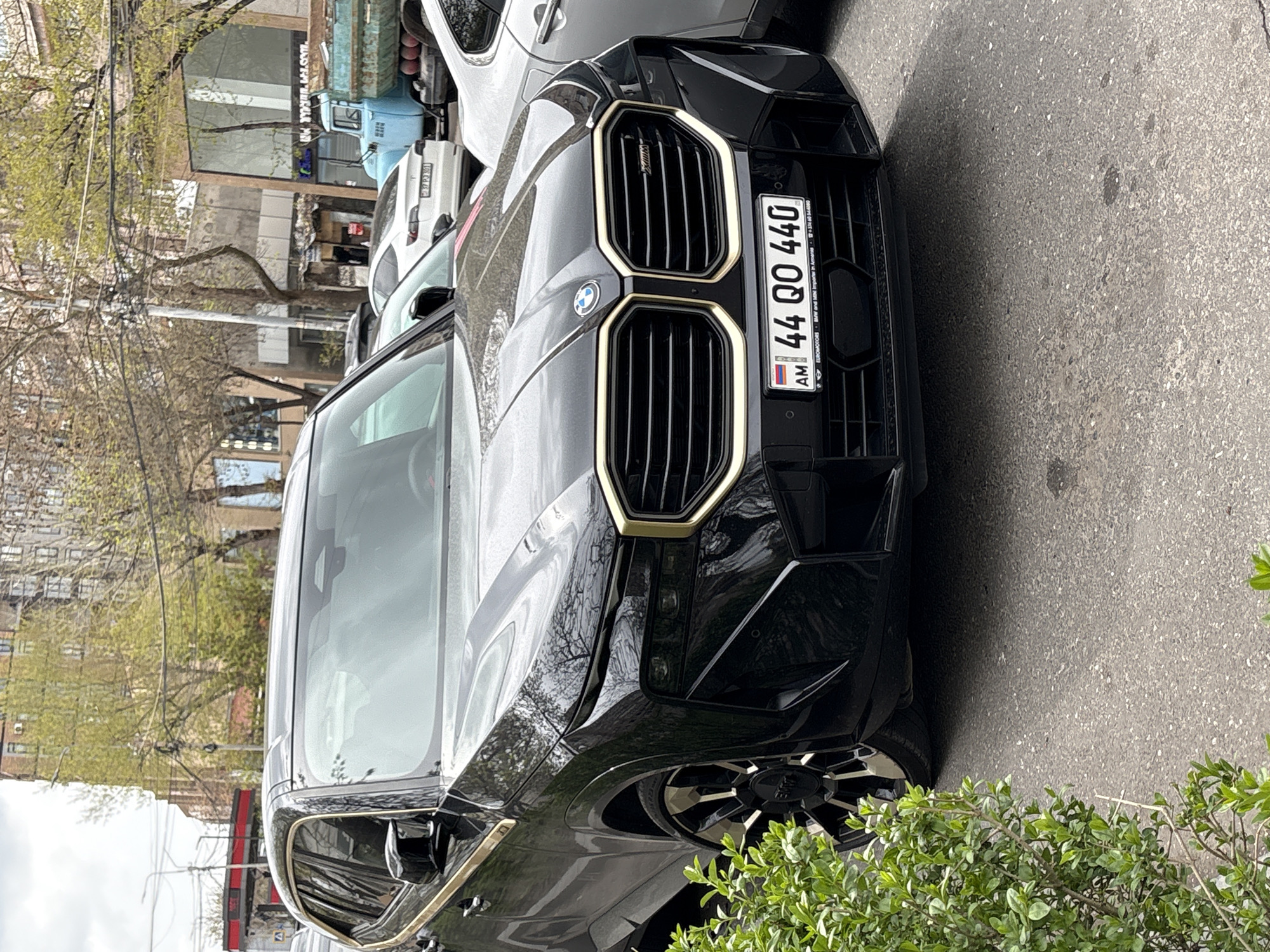 A black BMW, likely a newer model due to its design features, is parked on a city street. The car has a distinctive gold-trimmed grill and Armenia license plate number "44 QO 440" on the front. The vehicle appears to be in Yerevan, Armenia based on the license plate information. The background shows other parked vehicles, a building, and trees. A green bush is visible on the lower right side of the image, partially obstructing the view of the car.