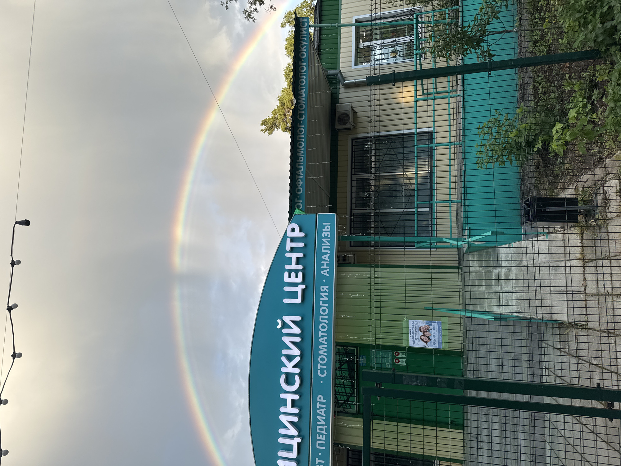 A full-frame, eye-level, outdoor, slightly low-angle shot captures a medical center in Bykovo, Russia, under a dramatic sky featuring a prominent rainbow. The building, painted in sections of teal and pale yellow, has large white lettering on its teal facade that reads "МЕДИЦИНСКИЙ ЦЕНТР" (Medical Center). Below this, additional text lists services: "• ПЕДИАТР • СТОМАТОЛОГИЯ • АНАЛИЗЫ" (Pediatrician, Dentistry, Analyses). To the right, a teal awning extends over the entrance, with more white lettering listing medical specializations: "• ОФТАЛЬМОЛОГ• СТОМАТОЛОГ• ОКУЛИСТ" (Ophthalmologist, Dentist, Oculist). The building features barred windows and an external air conditioning unit. In front of the building, a metal fence with teal accents encloses the property. A poster is affixed to the fence, depicting a smiling family with the text "АКЦИЯ ЗДОРОВАЯ УЛЫБКА - ЛУЧШЕ ВСЕГО!" (Promotion Healthy Smile - Best of All!). Concrete stairs and a ramp lead to the building's entrance. The ground in front of the fence consists of grey paving stones and some sparse greenery. The sky is overcast with a bright rainbow arching across it, suggesting recent rainfall. A string of lights is visible at the top of the frame. No people are present in the scene.