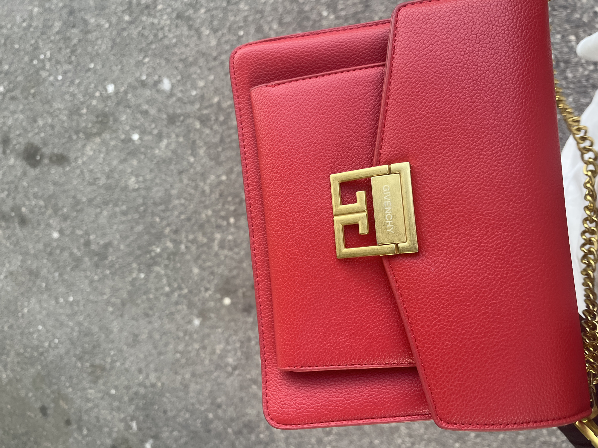 A close-up shot shows a bright red leather handbag with a gold clasp that reads "GIVENCHY". The background is a textured grey surface, likely asphalt. The handbag appears to be resting on the ground. No people or specific city details are visible.