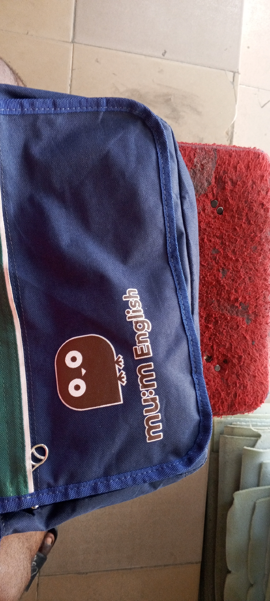 The image shows a close-up of a blue bag with a cartoon owl design and the text "mu:m English" printed on it. The bag has green and white stripes on the left side. To the right of the bag is a rough, red object, possibly a mat or a cushion, with dark speckles. The background consists of a tiled floor. The lower left corner of the image shows a person's foot wearing a black sandal. The scene appears to be indoors, possibly in a residential or commercial setting in Jalingo, Nigeria.