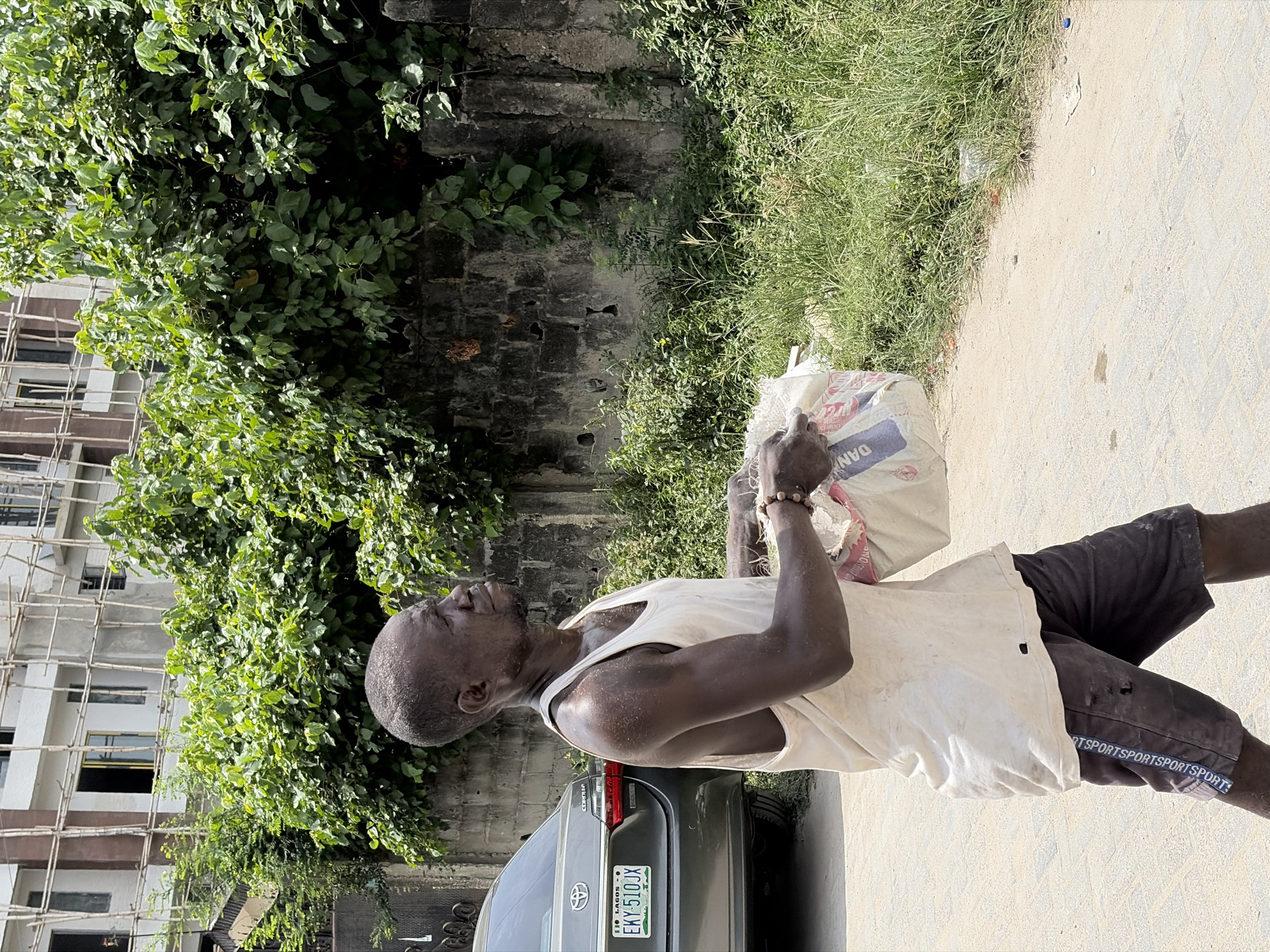 The image captures a man in Igboefon, Nigeria, carrying a sack. He is wearing a white tank top and dark shorts with "SPORTS" repeated along the side. A silver Toyota Corolla with the license plate "EKY-510JX" is partially visible behind him. The surroundings include a building under construction with scaffolding, a wall overgrown with greenery, and a paved surface. The scene appears to be outdoors, with ample sunlight.