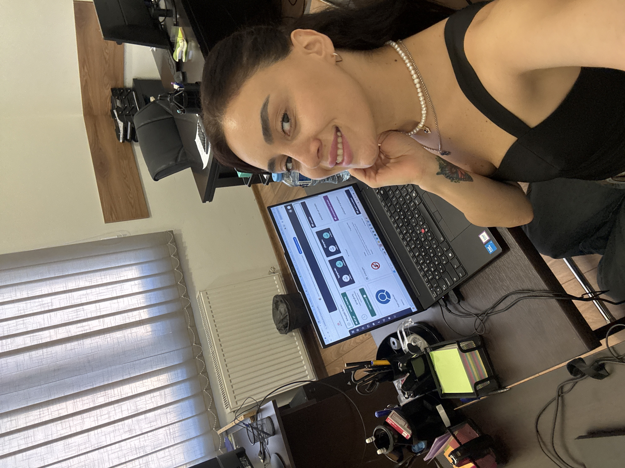 In a well-lit office space in Tbilisi, Georgia, a woman with dark hair is taking a selfie from a high-angle perspective. She is wearing a black top and a pearl necklace. Her left hand is resting on her chin, and she is smiling at the camera. In the foreground, her laptop is open and displays a website with Georgian text. To the left of the laptop, a desk organizer holds pens and scissors, and a stack of colorful sticky notes is visible. A radiator runs along the wall behind the desk, and vertical blinds cover the window. In the background, an office chair and another desk with computer monitors can be seen. The overall impression is of a professional workspace.