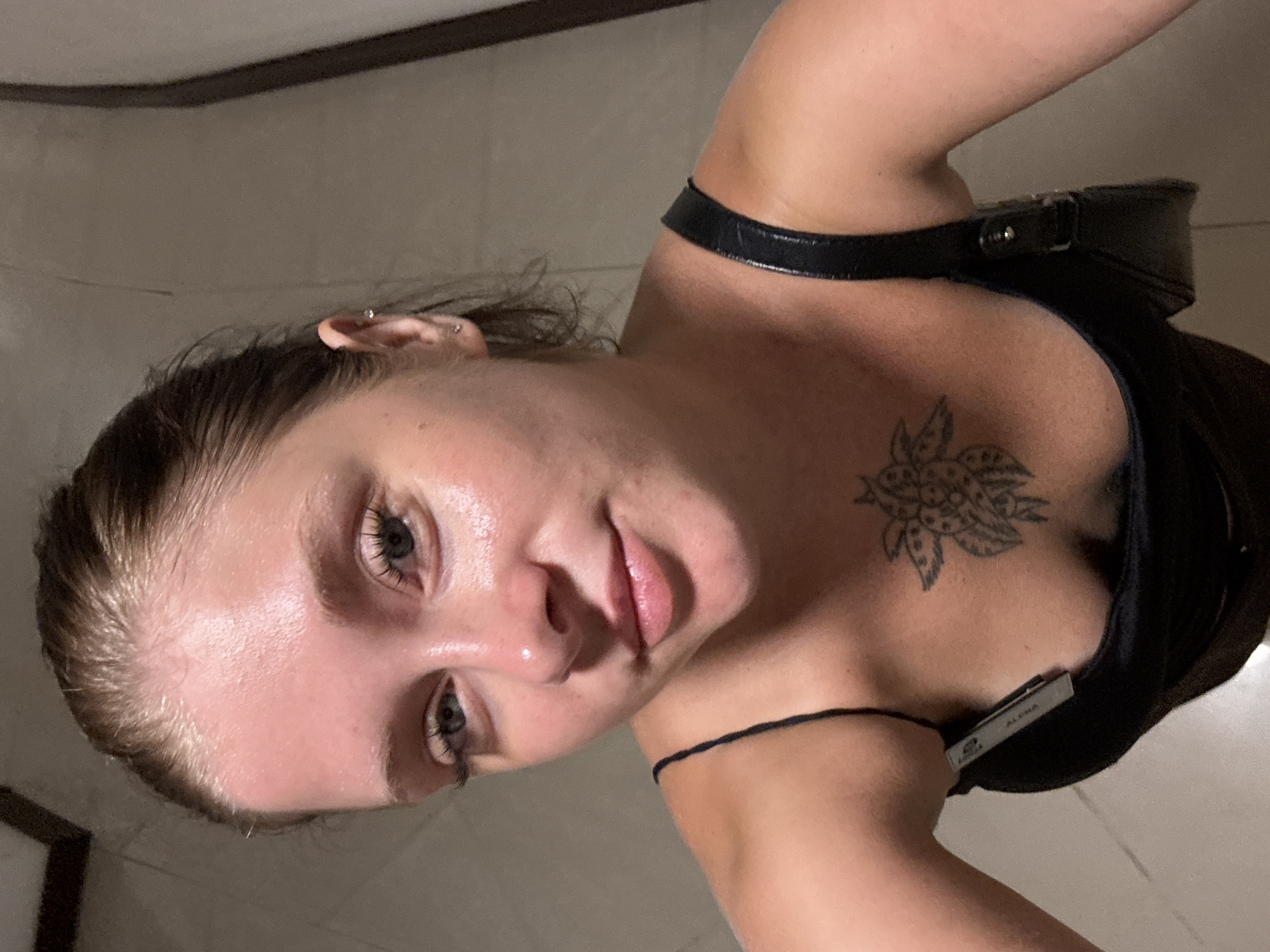 A woman with light skin and brown hair pulled back is shown in a close-up, downward-facing selfie. She is wearing a black tank top with thin straps. A black bag with a shiny strap is visible on her right shoulder. She has a tattoo of a flower on her chest. Her eyes are looking towards the camera, and her lips are slightly parted. The background appears to be an indoor setting with tiled walls. There are no visible landmarks or clues to indicate a specific location in Aksu, Turkey.