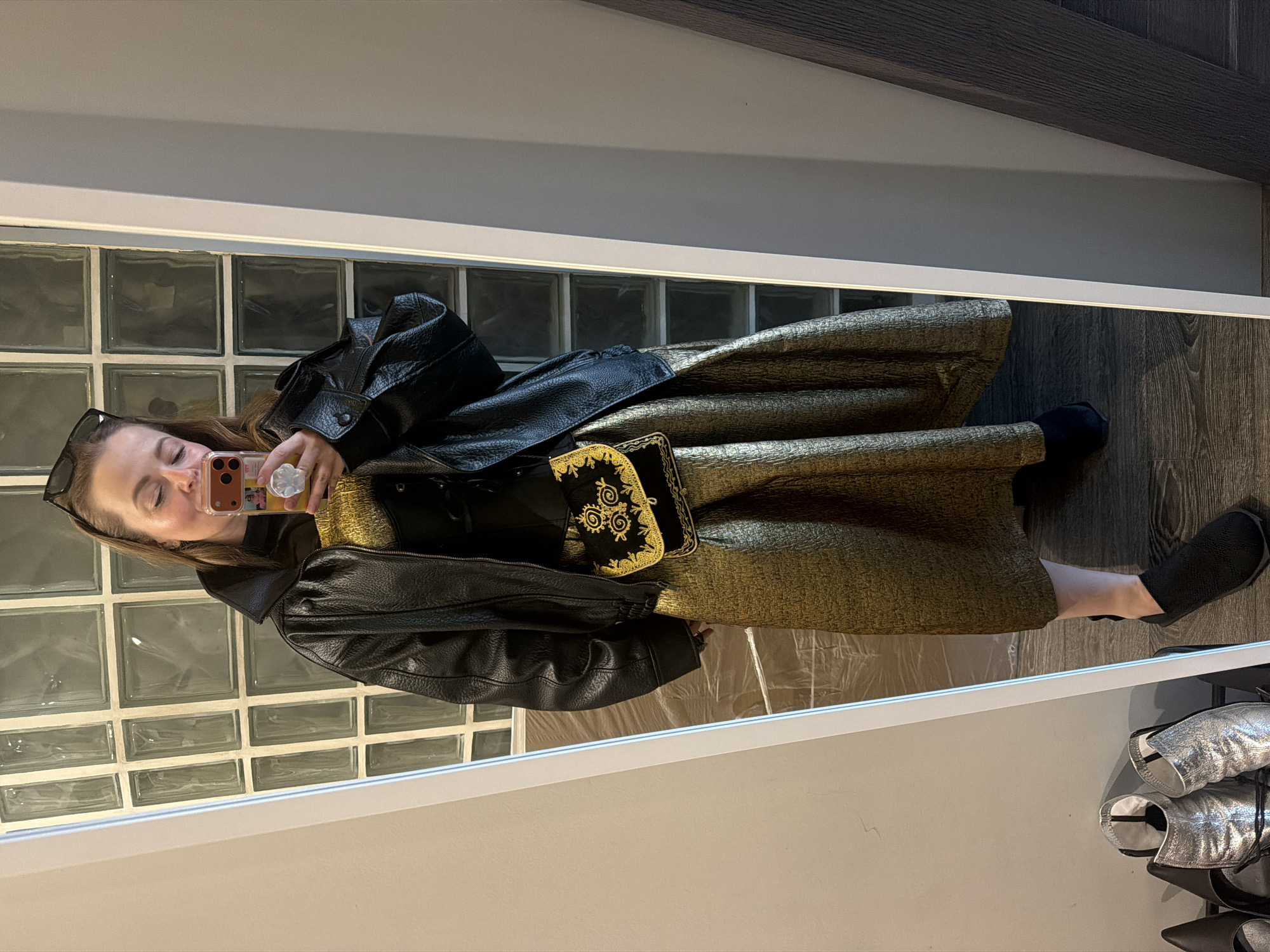 The image shows a woman taking a selfie in front of a mirror. She is wearing a gold dress, a black corset, a black leather jacket, and black shoes. She is holding a cell phone with an orange case and a white flower-shaped grip. Sunglasses are perched on top of her head. A black purse with gold embroidery is visible. The background includes a wall of glass blocks and a shoe rack with silver shoes. The location is in the city of Unknown, Unknown.