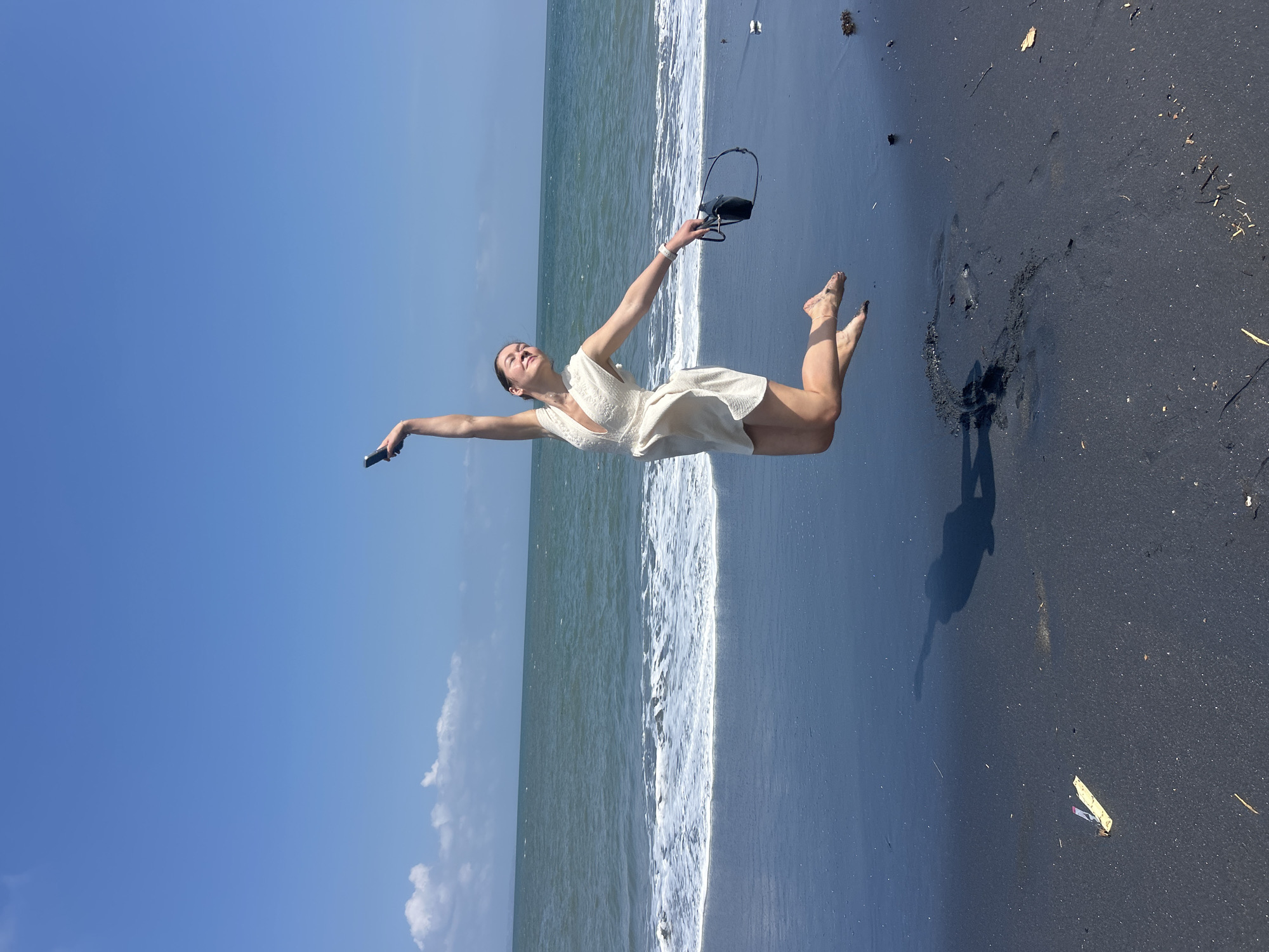 A young woman is captured mid-air, leaping joyfully on a black sand beach. She wears a short, white, V-neck dress and her feet are bare. Her left arm is raised, holding a dark-colored smartphone aloft, while her right arm is extended, clutching a dark pair of sunglasses. The ocean stretches out behind her, with gentle waves breaking on the shore under a clear blue sky dotted with a few white clouds. The black sand is wet near the water's edge, reflecting the sky and the woman's shadow, which is cast prominently on the ground. The location is identified as the city of Mengwi, Indonesia.
