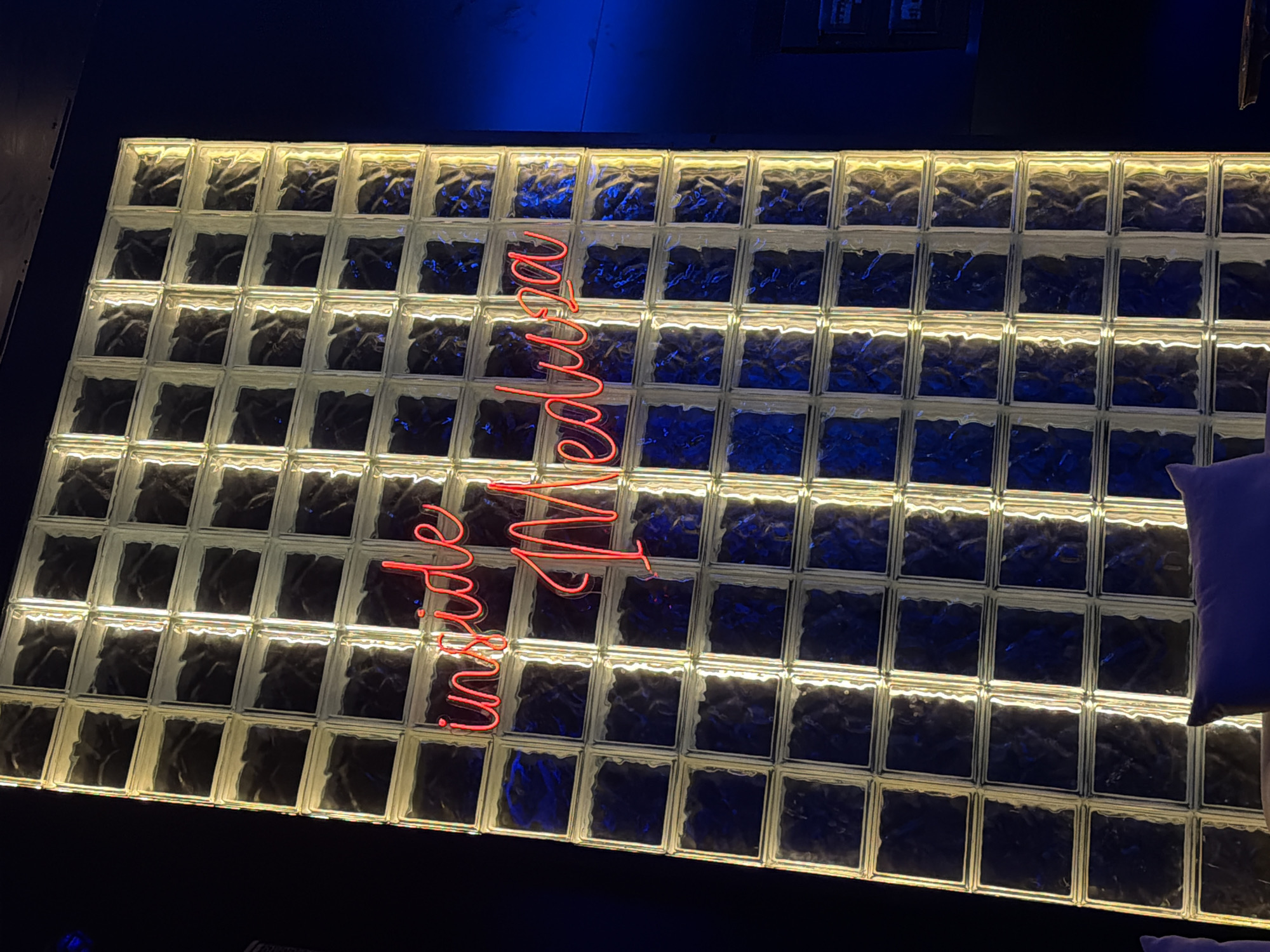 The image shows a wall constructed of glass blocks, illuminated from behind, creating a grid pattern with a warm, yellowish glow. A neon sign with red cursive lettering reads "inside Meduza". The sign is positioned in the center of the wall, spanning across multiple glass blocks. The background is dark, with a hint of blue lighting on the right side, suggesting an interior space, possibly a bar or club. There are hints of furniture, specifically a purple cushion, in the foreground, indicating that the viewer is looking into a seating area. The overall impression is that of a stylish, possibly dimly lit establishment. This scene is located in Nha Trang Ward, Vietnam.