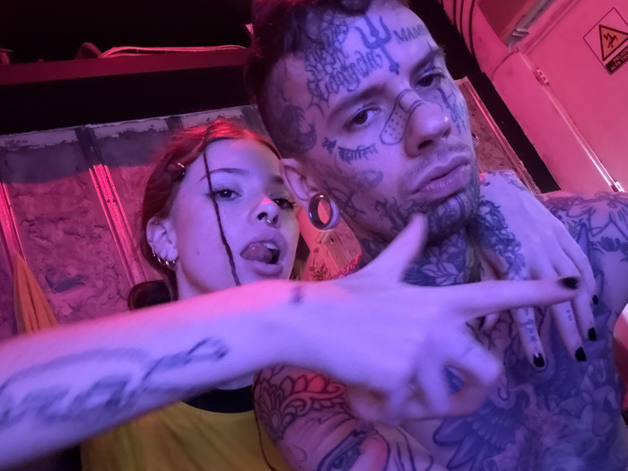 The image depicts two individuals, one male and one female, in a dimly lit indoor setting dominated by pink and magenta artificial lighting. Both subjects are positioned close to the camera, suggesting a selfie.
The male subject, on the right, is heavily tattooed across his face, neck, and upper torso. Notable facial features include a small bandage on his nasal bridge, large ear gauges, and various tattoos, including a trident symbol on his forehead and script. He is shirtless and has a beard and mustache.
The female subject, on the left, has reddish-brown hair with some strands pulled forward. She features multiple ear piercings and a nose piercing. Her tongue is partially extended, and she wears dark eyeliner. Her left arm is extended towards the camera, with her hand forming a "finger gun" gesture; her fingernails are painted black. Her right hand rests on the male subject's tattooed chest. She is wearing a yellow top with a dark collar.
The background reveals industrial-style elements. To the upper right, a light-colored structure displays a yellow and black warning sign, partially legible as "PELIGRO ALTA TENSIÓN" (Danger High Voltage). Further left, a dark, possibly corrugated surface is visible, and a yellow fabric item is partially seen behind the female subject.
The presence of the "PELIGRO ALTA TENSIÓN" sign indicates a Spanish-speaking environment, consistent with Buenos Aires, Argentina. However, a more specific location within the city cannot be determined from the visual information provided. The scene suggests an entertainment venue or a converted industrial space.