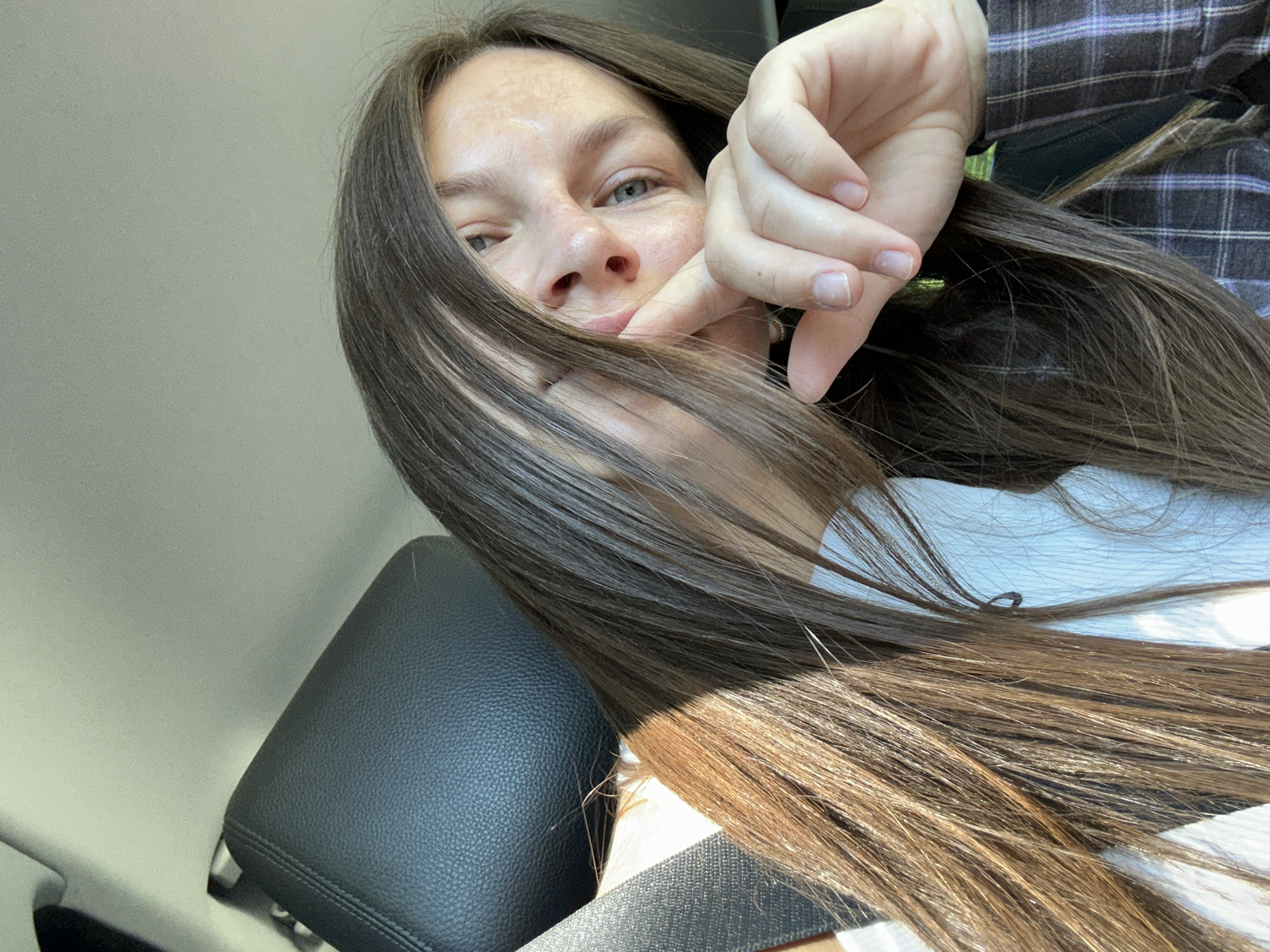 A young woman with long, dark brown hair is captured in a close-up, indoor selfie. She is seated in a vehicle, with a black leather car seat and a seatbelt visible in the background. The woman is looking at the camera, with her hand touching her chin and mouth. She is wearing a white top and a plaid jacket. The lighting suggests it is daytime, with sunlight casting shadows on her hair and face. The location is identified as Jabuka, Montenegro.