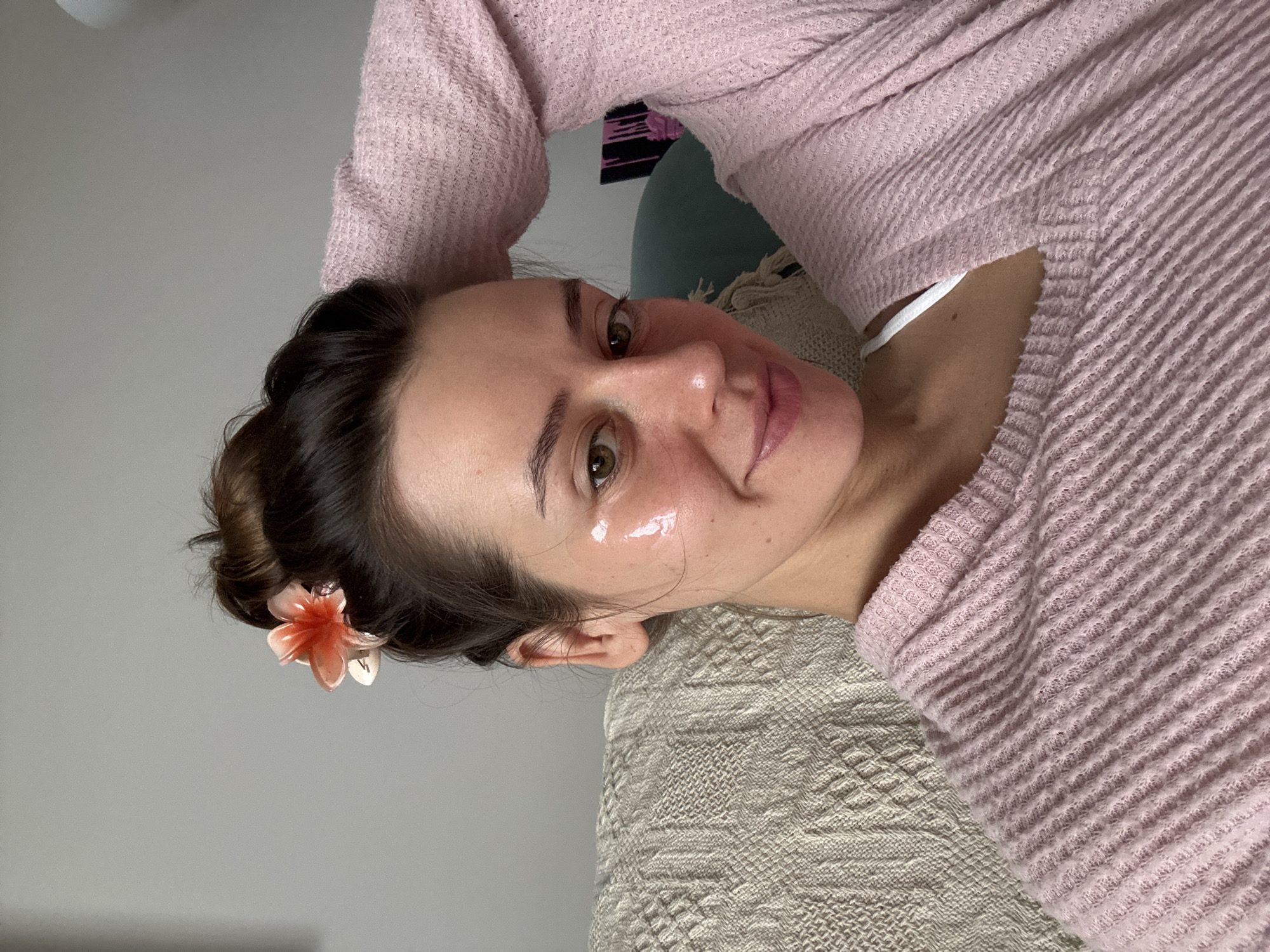This media file presents a diagonal, medium close-up portrait of a young woman resting indoors. The woman, with fair skin and dark brown hair styled in an updo, has a small, light pink and orange flower clip in her hair. She possesses hazel eyes, dark eyebrows, and her lips are tinted with a light pink/mauve color. Her cheeks show a glossy sheen, possibly from a skincare product like a face mask or moisturizer, suggesting a moment of self-care.
She is wearing a light pink, long-sleeved top with a visible ribbed or waffle-knit texture. A white strap, likely from an undergarment, is visible beneath her top near her shoulder. The woman is lying down, with her head resting on a light-colored, textured blanket or throw, which features a woven pattern in shades of beige or light grey.
She looks directly at the camera with a gentle, relaxed smile, conveying a calm and pleasant demeanor. The background is simple, featuring a plain light grey wall behind her head. A small, obscured detail of what appears to be a darker object with black and pink patterns is partially visible in the upper background, behind her pink top. The setting appears to be a casual, indoor environment. There is no visible text in the image. The contextual location is Krasnoyarsk, Russia.