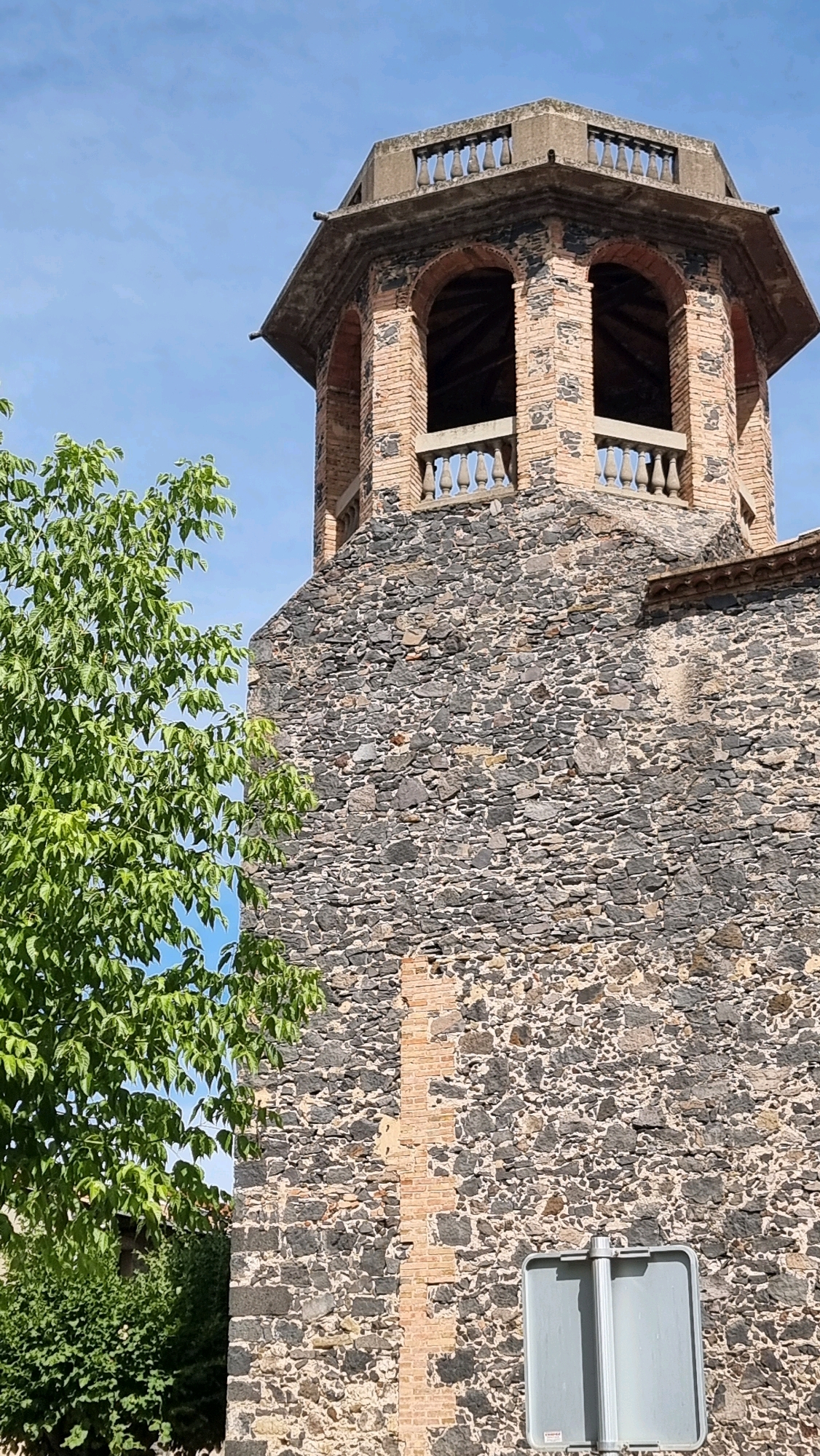 The image shows the bell tower of a stone building in Castellfollit de la Roca, Spain. The tower has a section made of dark gray stones and another section made of reddish-brown bricks, with arched openings and a concrete roof. A green tree with lush leaves is positioned to the left of the tower, partially obscuring the view of the lower part of the building. A grey sign is visible on a pole at the bottom right of the image. The sky is clear and blue.