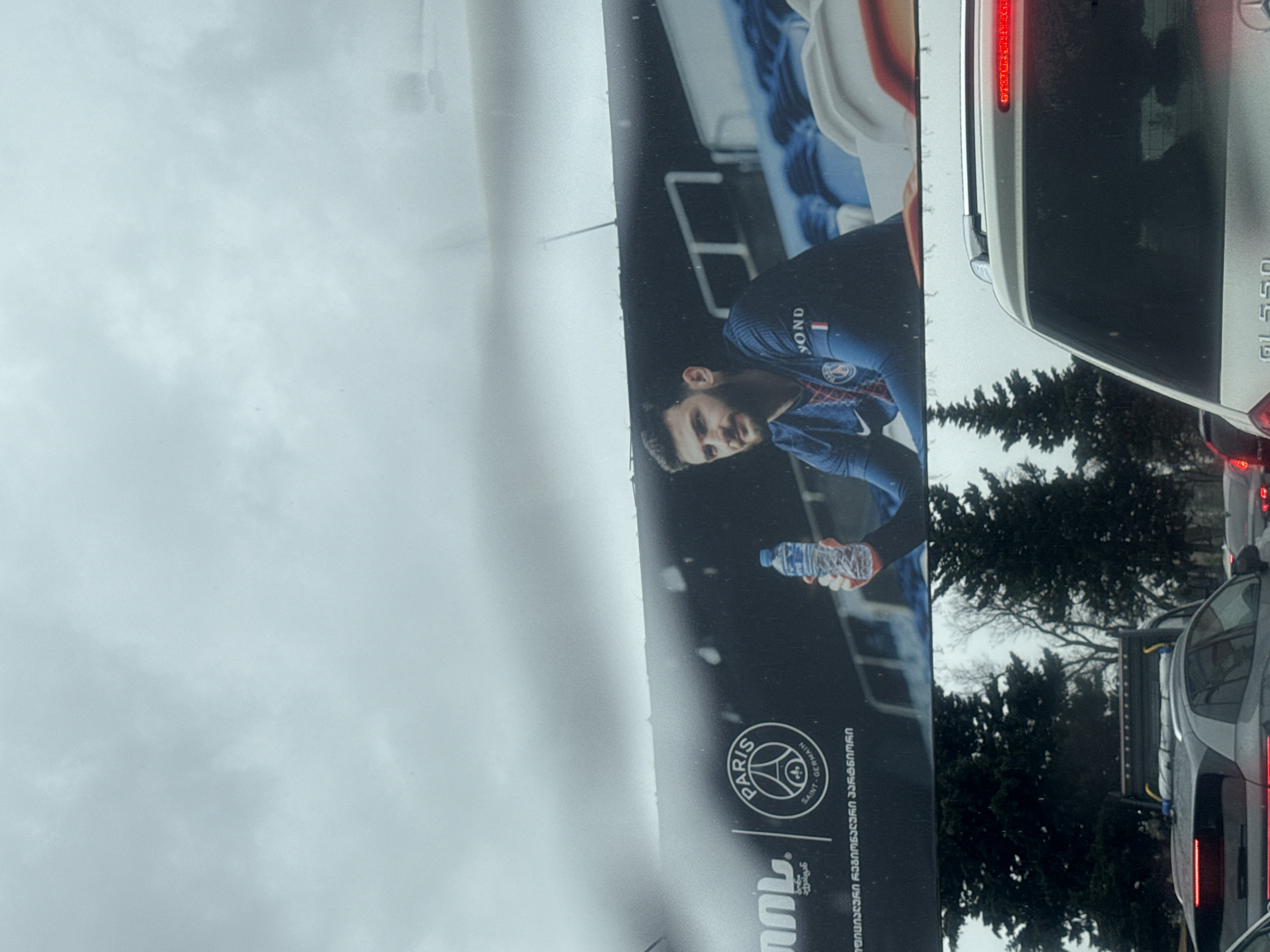 The image depicts a street scene in Tbilisi, Georgia on a cloudy day. A billboard advertisement for Paris Saint-Germain (PSG) featuring a soccer player holding a water bottle is visible. The billboard includes the PSG logo and text in Georgian. Cars are in traffic on the road, including a white Mercedes-Benz GLS 550. Evergreen trees are in the background.
