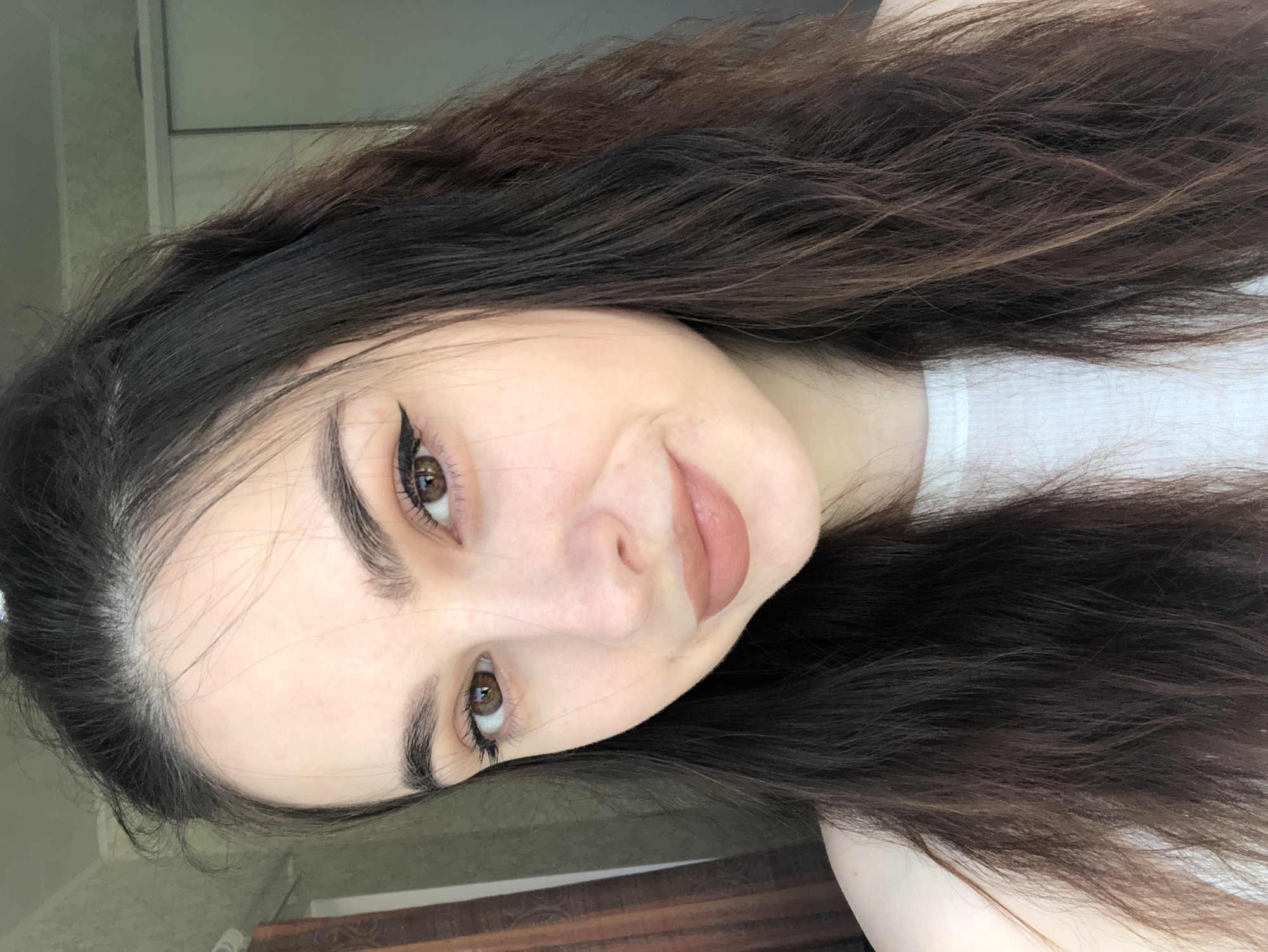 A young woman with long, wavy dark brown hair is posing for a selfie. She has fair skin, brown eyes with winged eyeliner, and light brown lipstick. She is wearing a white shirt. The background includes a light-colored wall or door and a glimpse of a wooden surface. There's no indication of the location being Noviy, Russia.