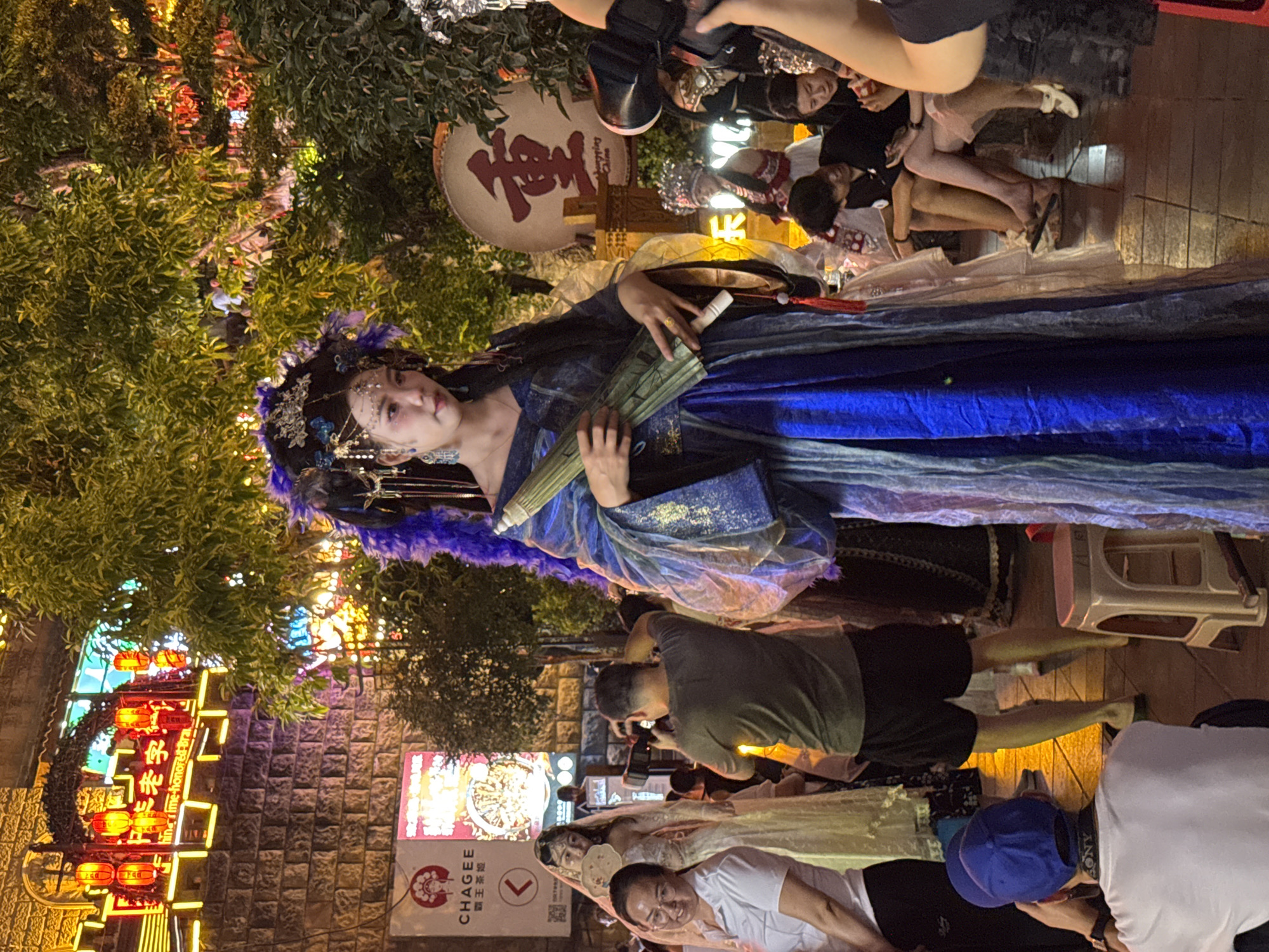 The image depicts a nighttime scene on a street, likely in Yuzhong District, China, illuminated by neon signs and string lights. In the foreground, a woman in elaborate traditional Chinese attire, including a blue and gold robe, a headdress with purple feathers, and intricate jewelry, holds a light-colored umbrella. She is facing slightly to her left, looking at the camera with a neutral expression.
Behind her, a man in a green t-shirt and black shorts is taking a photo with a camera. To the left of the main subject, another woman in a white shirt and glasses smiles at the camera. Further back, several other people are visible, some seated and others standing, also observing the scene. A sign with Chinese characters and the word "CHAGEE" is visible on a building to the left. A large circular drum with Chinese characters, possibly spelling "Chongqing China", is mounted on a post in the background. The environment appears to be a pedestrian area with stone-paved ground and trees with lit foliage. The overall atmosphere suggests a cultural event or performance.
