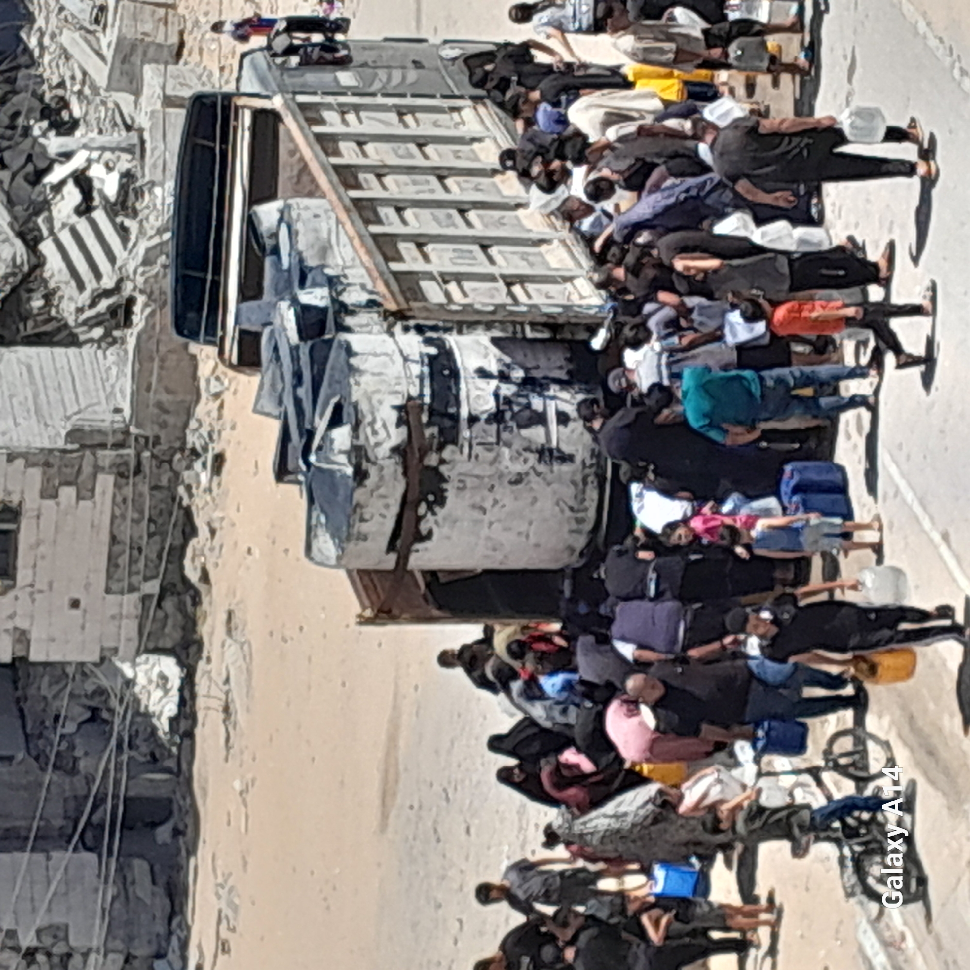 In Deir el-Balah, Palestinian Territories, a large group of people are gathered around a truck laden with large plastic water tanks. The truck is parked on a dirt road, with the backdrop of damaged buildings indicating a recent conflict or destruction. Many individuals are carrying smaller containers, some of which appear to be jerrycans or water bottles, suggesting they are collecting or distributing water. The scene depicts a humanitarian effort, likely related to water scarcity or providing essential supplies to the community. Some people are standing by the truck, while others are moving through the crowd, interacting with each other and managing the containers. The environment is dusty and appears to be a densely populated area. A young boy is also visible riding a bicycle in the foreground.