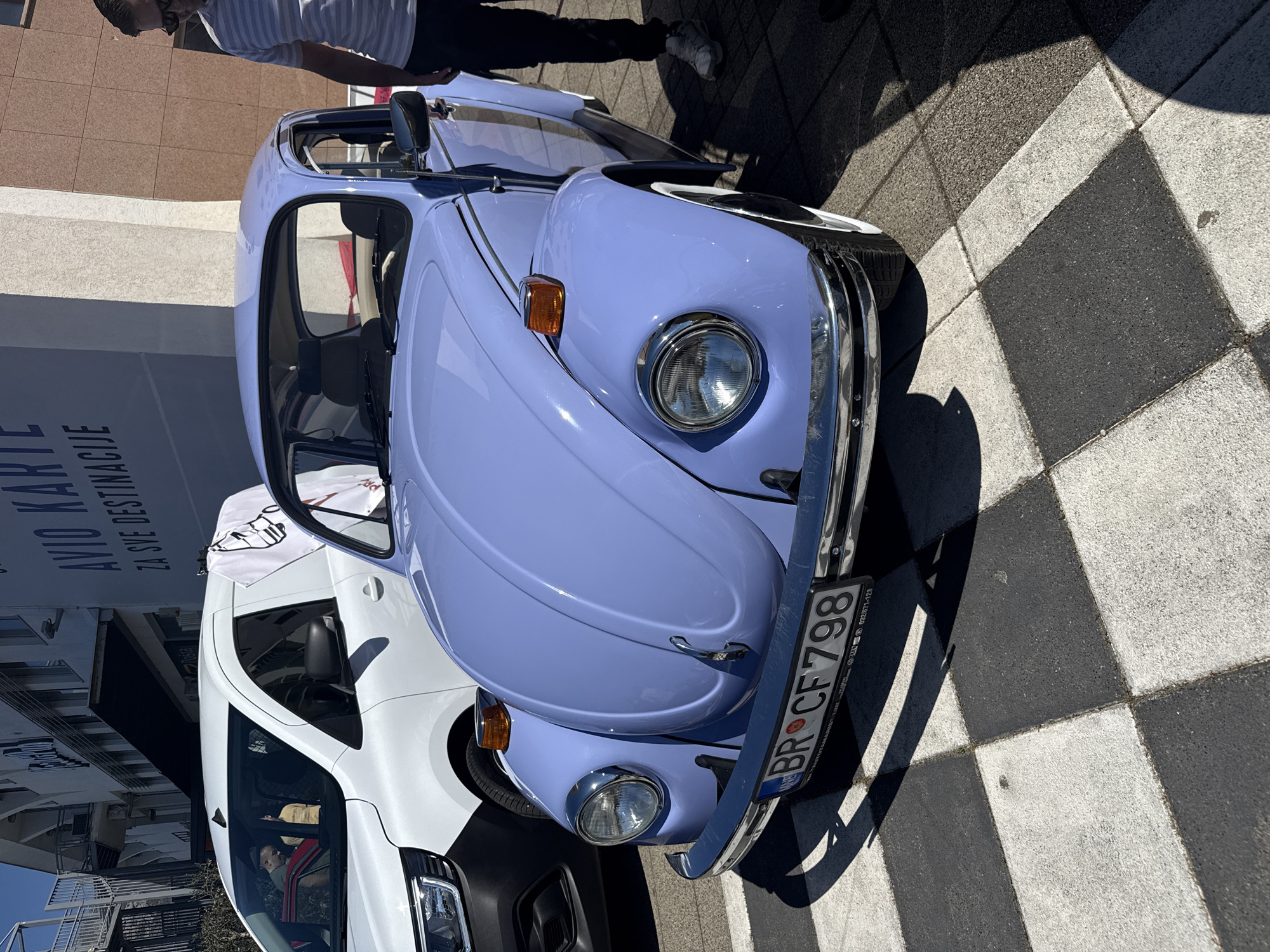 The image features a periwinkle blue Volkswagen Beetle parked on a checkered sidewalk. The license plate reads "BR CF 798". A white van with a partially visible driver is parked behind the Beetle. A building with the partial text "AVIO KARTE ZA SVE DESTINACIJE" is in the background. The photo appears to be taken outdoors on a sunny day in a city.