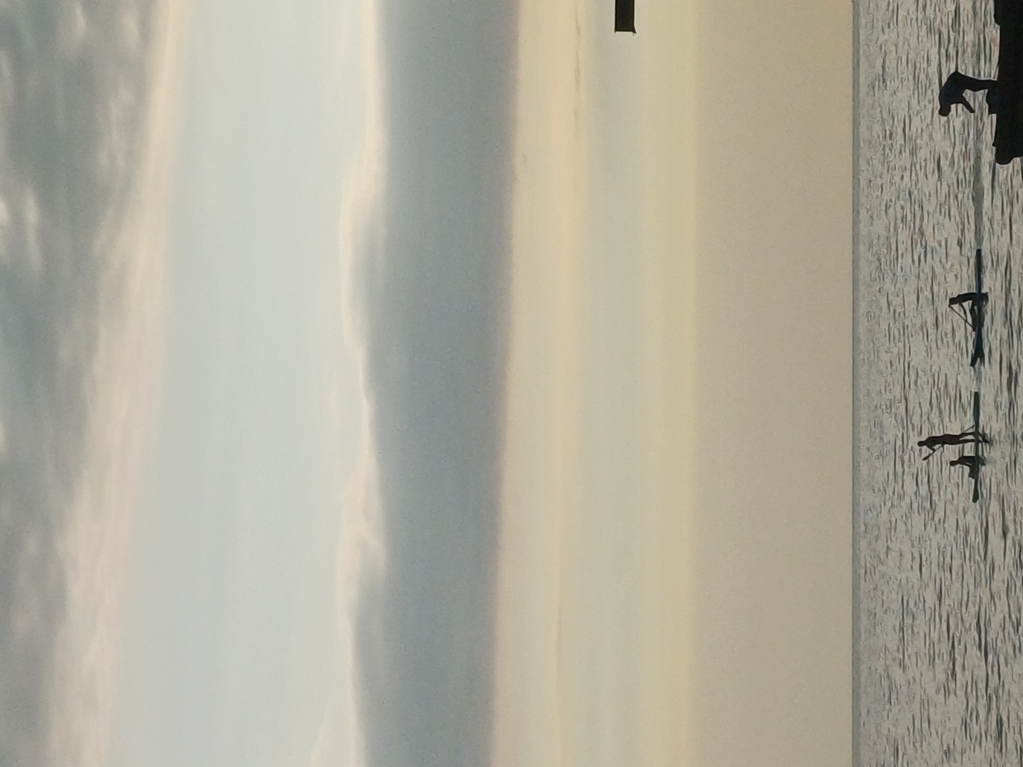 The image depicts a serene scene of paddleboarders on the Black Sea, likely near the city of Sochi, Russia, given the visual context. The sky is overcast with distinct horizontal cloud formations. The sea is calm, with gentle waves. In the foreground, several individuals are engaged in paddleboarding. One paddleboarder stands, holding a paddle, while another is seated on a paddleboard. Further to the right, another individual stands on what appears to be a dock or a breakwater, with a paddleboard nearby. The overall lighting suggests either early morning or late evening, with soft, diffused light. The silhouettes of the people and the paddleboards are clearly visible against the lighter backdrop of the sea and sky.