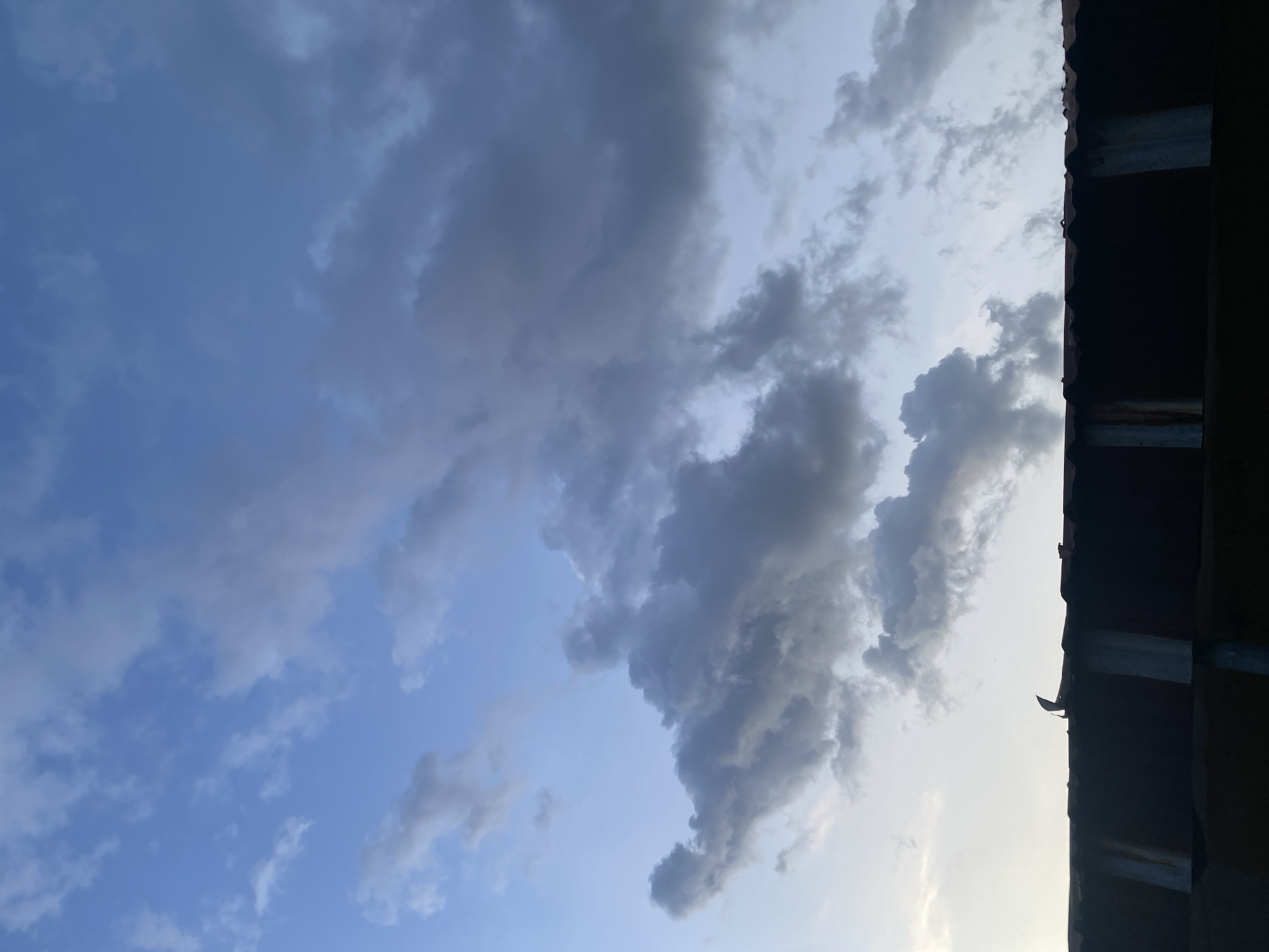 The image displays a sky scene captured in portrait orientation, with a section of a building's roofline silhouetted along the right side. The sky is largely filled with clouds against a varied blue backdrop.
The clouds range from scattered, lighter white and grey wisps in the upper left to denser, darker grey formations dominating the central and lower right portions of the sky. These heavier clouds suggest changing weather conditions, possibly indicating a build-up before rain or a generally overcast period. The sky's blue varies in intensity, hinting at either early morning or late afternoon light, with some areas appearing brighter than others, implying the sun's position might be just outside the frame or behind the clouds.
The structure on the right is a dark, almost black, silhouette, appearing to be the roofline of a building. It extends vertically through the entire right edge of the frame. Visible details suggest it is constructed from corrugated metal sheets, with what appear to be support beams or brackets underneath, showing some signs of rust or wear. A small, distinct dark object, possibly debris or a part of the roof fixture, is discernible near the top of the roofline.
The image captures a static moment of the sky and a building, characteristic of an urban or semi-urban setting like Jos, Nigeria. No people, specific activities, or visible text are present. The scene primarily showcases the natural beauty and dynamic elements of the sky and weather.