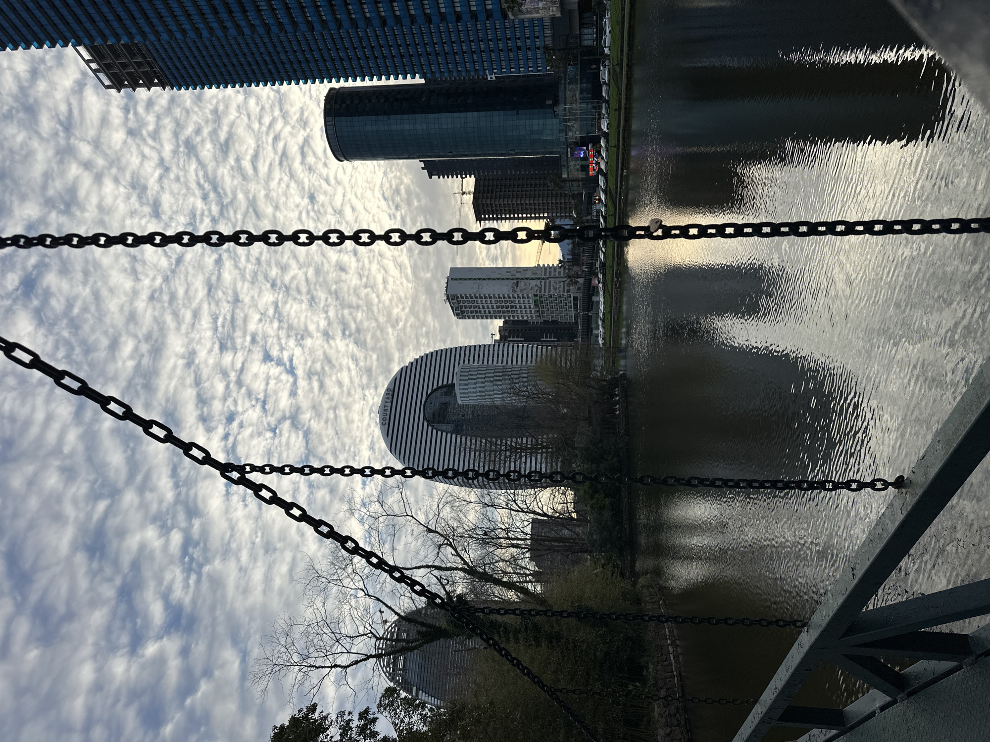 The image is taken from a low angle, looking upwards at modern architecture and a river in Batumi, Georgia. A metal chain and part of a bridge structure are in the foreground, partially obscuring the view.
In the background, several tall, modern buildings are visible. One building has a distinctive curved facade with alternating black and white stripes. The river reflects the buildings and the sky, which is filled with numerous small, puffy clouds. Some trees with bare branches are also present, adding to the urban-nature mix.