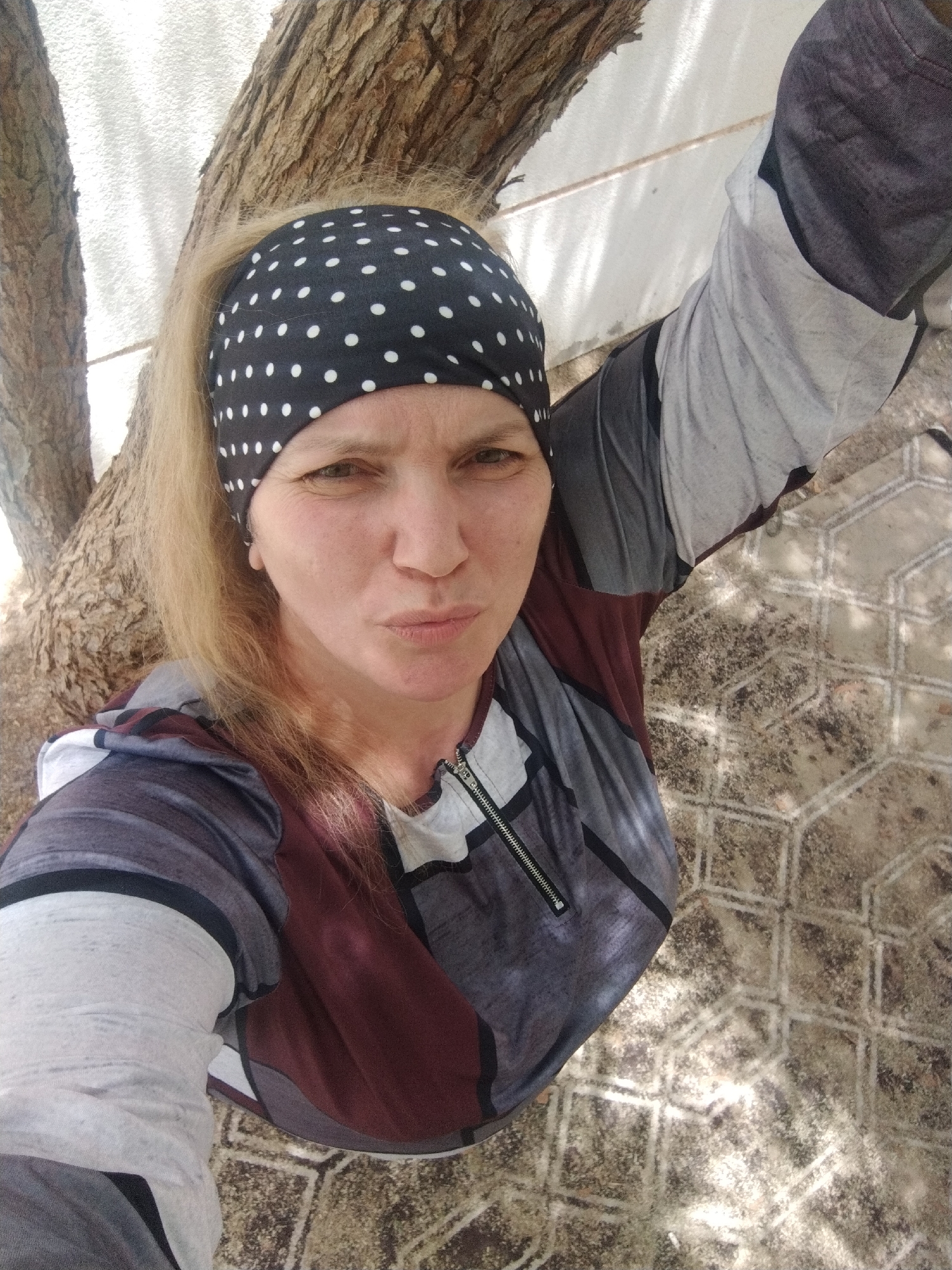 A woman with blonde hair and a black and white polka dot headscarf stands next to a tree. She is wearing a multi-colored zippered top. The ground is paved with hexagonal tiles. The location is Al Mas'oudi, United Arab Emirates.