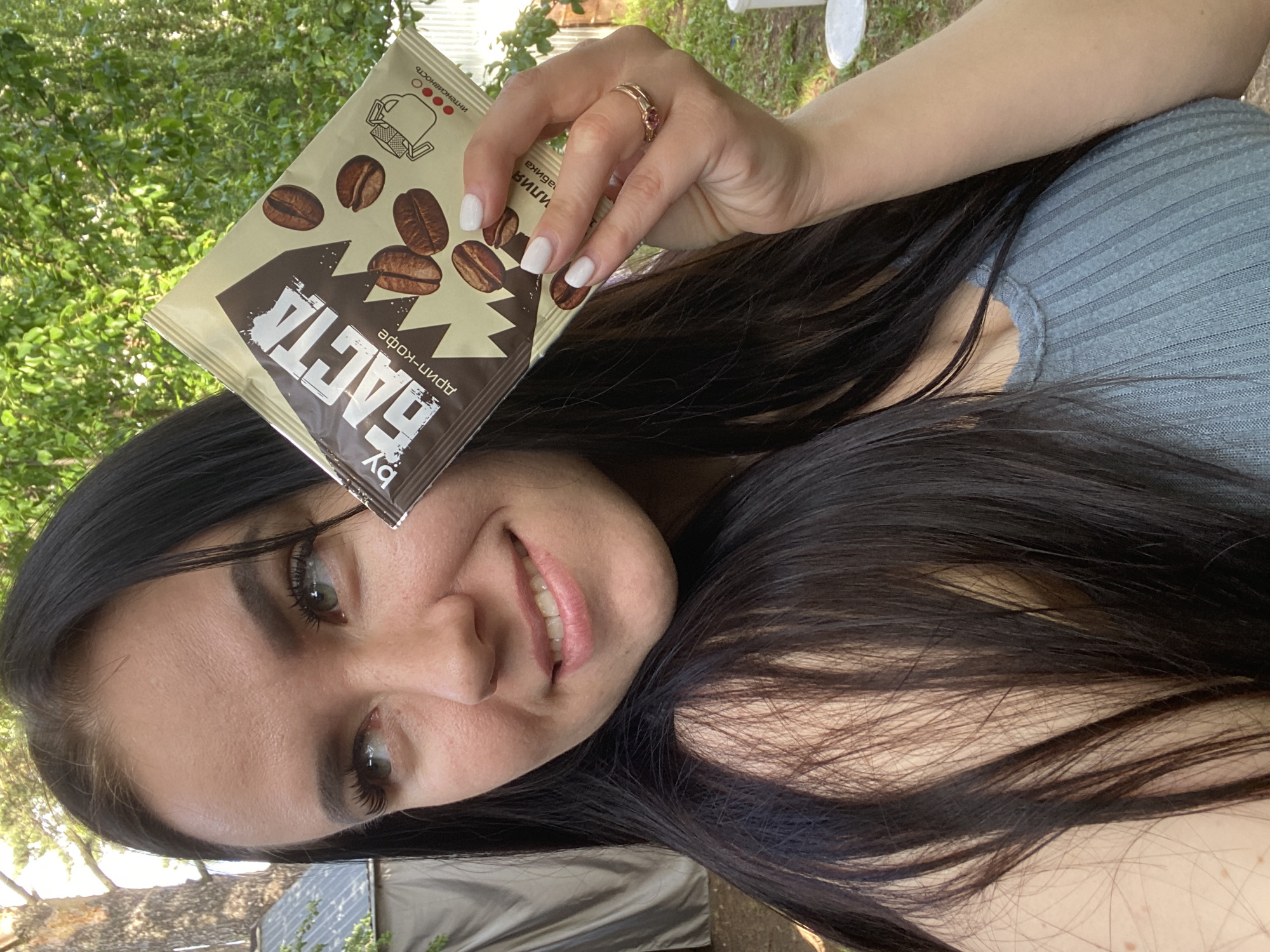A woman is holding a packet of "BASTA" brand coffee in an outdoor setting. The woman has long, dark hair, fair skin, and is wearing a light blue ribbed top. She is smiling slightly. The coffee packet has coffee bean illustrations and text in Russian. The background shows green foliage and a hint of a white object (possibly a chair). The image seems to be taken during daylight. There is no information in the image itself to confirm it's from the city of Chaikovsky, Russia.