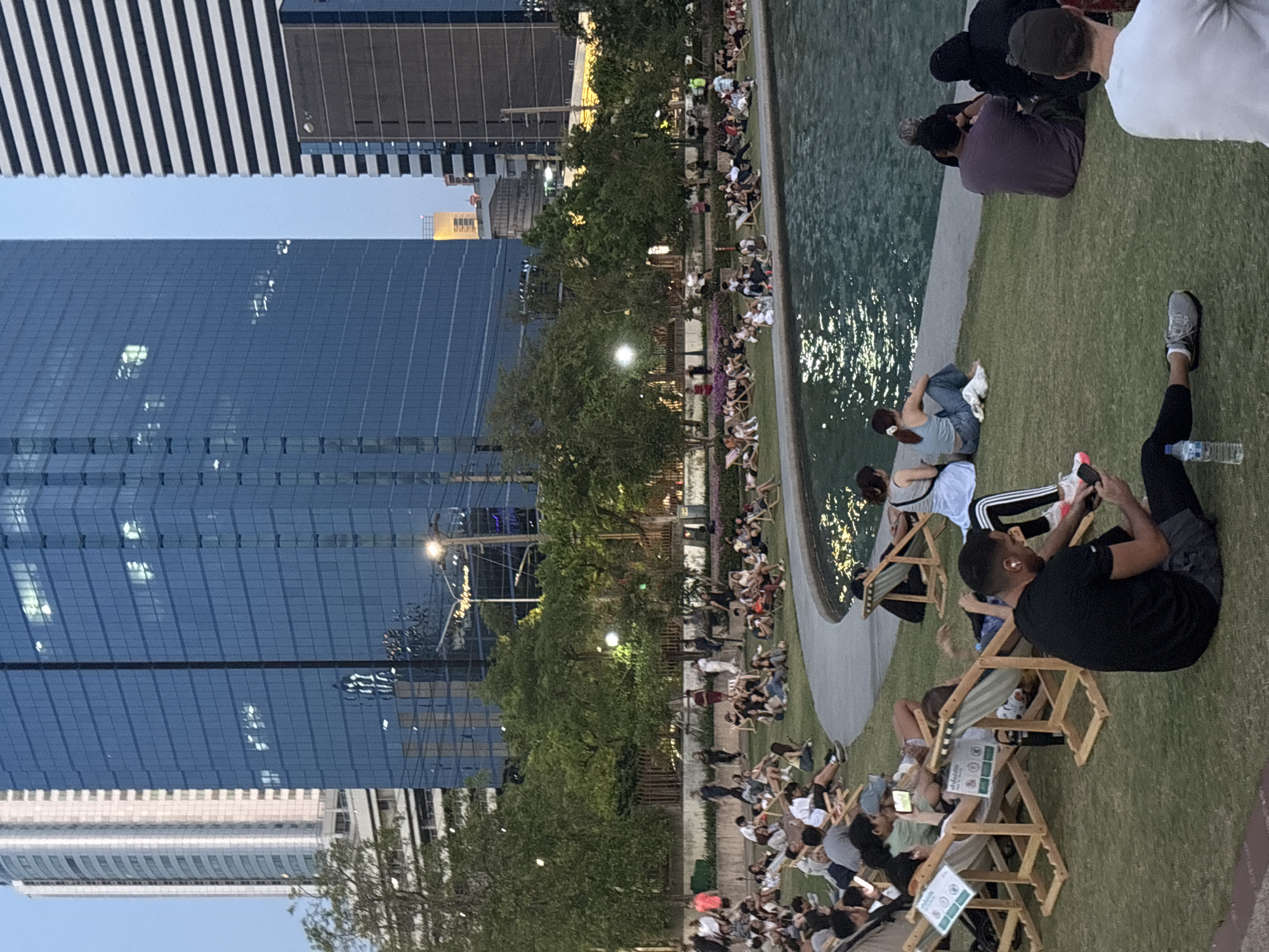 The image depicts an outdoor gathering in Bangkok, Thailand, with a cityscape backdrop dominated by modern skyscrapers. A group of people is seated on folding wooden chairs or directly on the grass alongside a water feature, possibly a pond or fountain. Many individuals are engaged with their mobile devices. The scene takes place in a public space, likely a park or plaza, during twilight or early evening. The atmosphere appears relaxed and social.