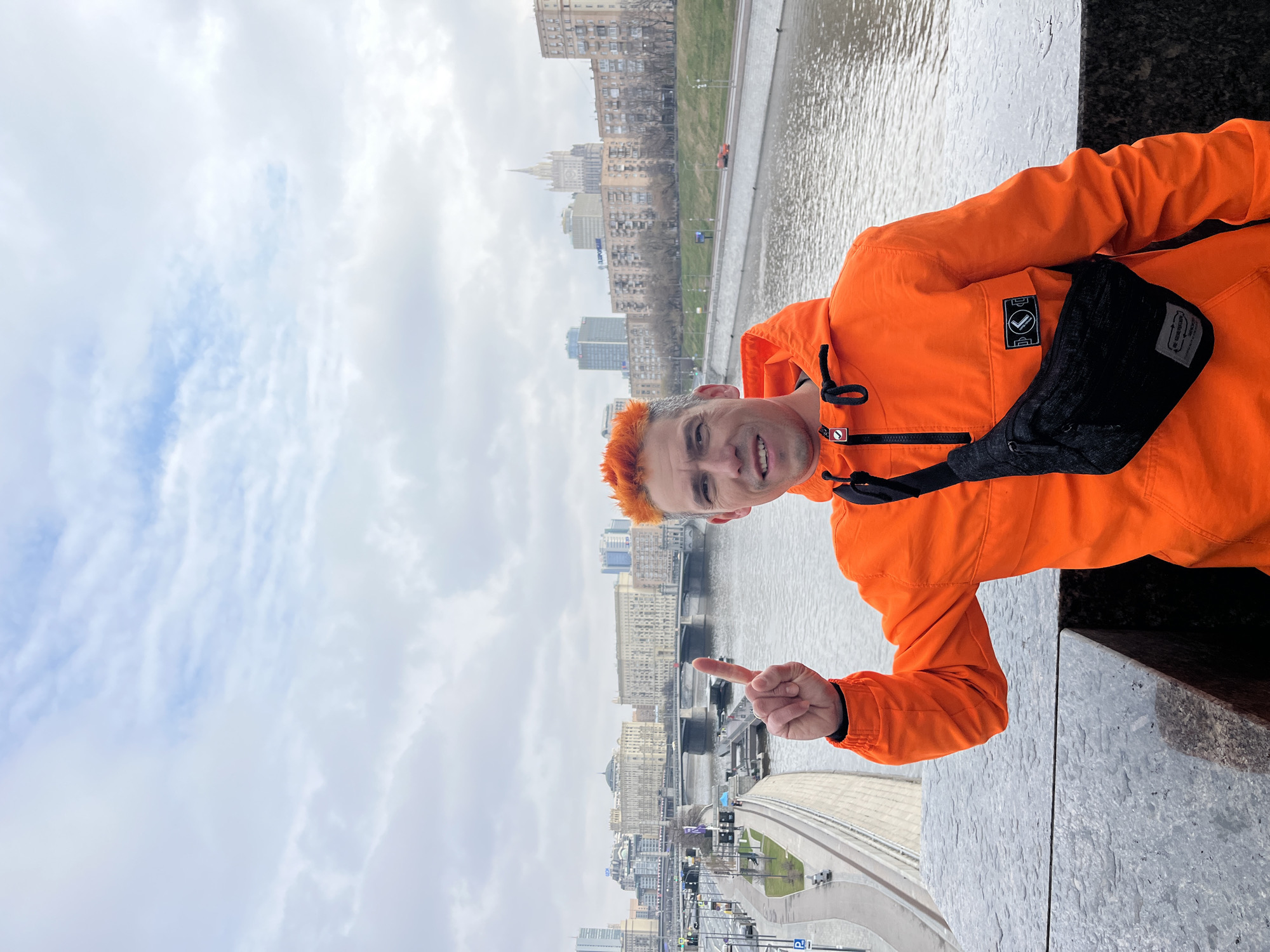 A man with orange hair is standing on a high platform, possibly a bridge or building, with a river and city skyline in the background. He is wearing an orange jacket and a black crossbody bag, and is pointing his finger towards the cityscape. The city features a mix of old and modern buildings, and the river has visible water flow. The sky is overcast with cloudy conditions. The man appears to be taking a selfie or posing for a picture.