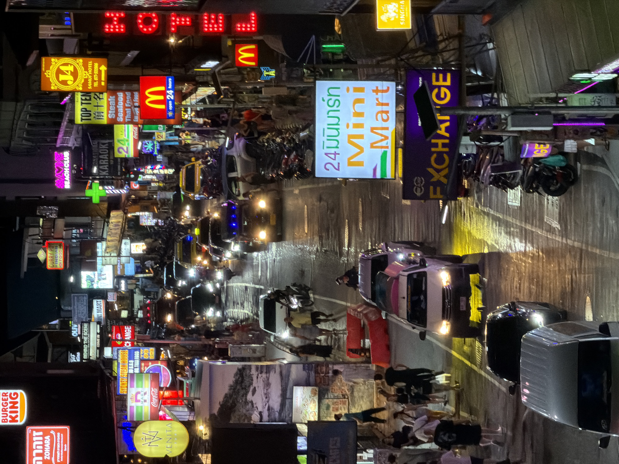 The image depicts a bustling street scene at night in Ban Rak, Thailand. The street is lined with numerous neon signs advertising various businesses, including Burger King, Zohara, Olive, Mini Mart, McDonald's, and a "HOTEL." Several cars and motorcycles are visible on the wet street, suggesting recent rain. Pedestrians are walking along the sidewalks, some of whom are illuminated by the neon lights. The atmosphere appears lively and commercial.
