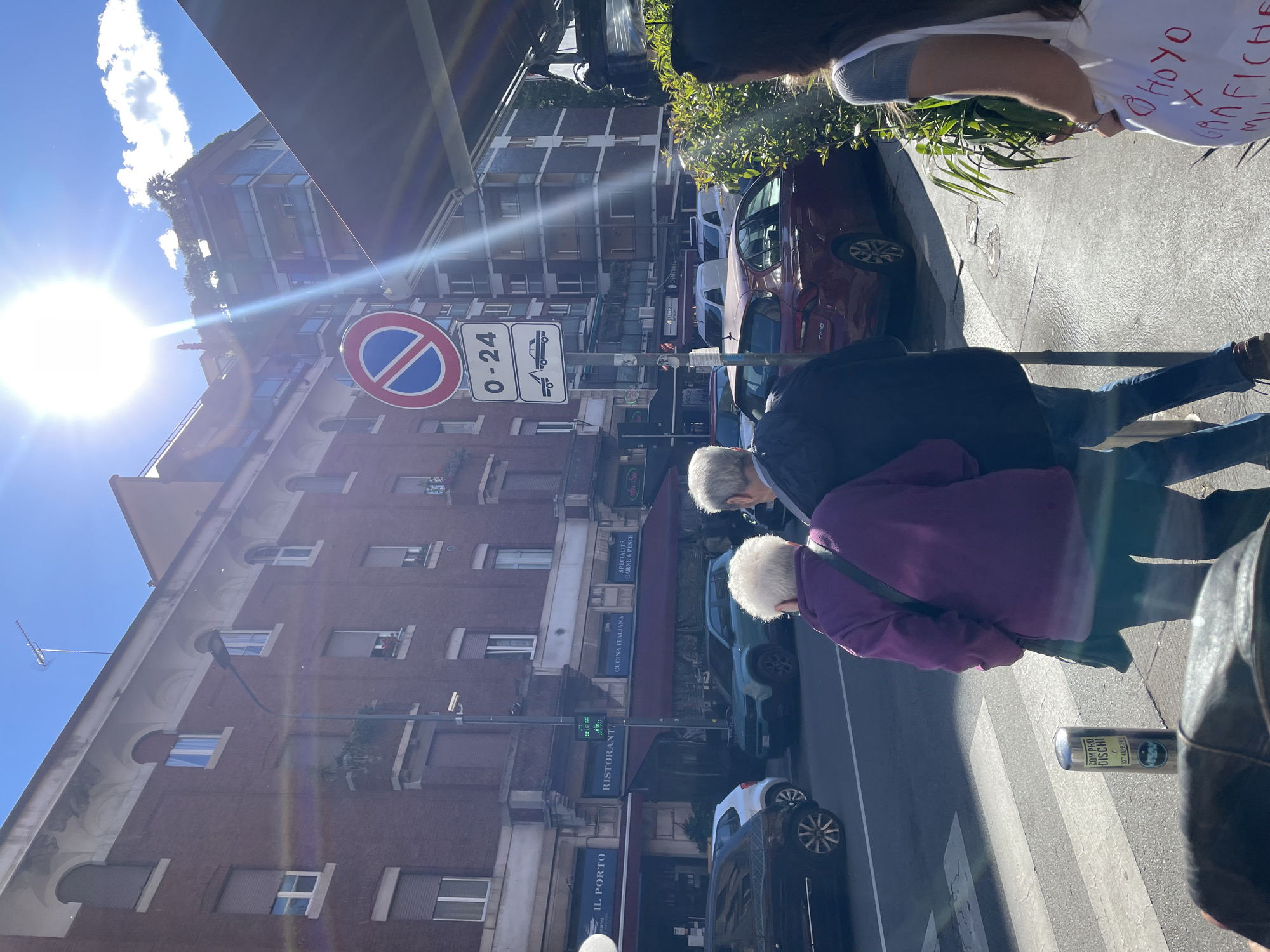 The image captures a street scene in Milan, Italy, evident from the signage and architectural style. A couple with white hair are crossing a zebra crossing in the foreground. A pole with the text "COMPRO DISCHI" (I buy discs) is visible, with a phone number. In the background, there is a brown brick building with multiple windows and a sign displaying "IL PORTO RISTORANTE" (The Port Restaurant) with "CUCINA ITALIANA SPECIALITA CARNE & PESCE" (Italian Cuisine Specialities Meat & Fish). Above that is a sign with Italian "no parking" and "towing" symbols and "0-24" (hours). Several cars are parked along the street. The sun is shining brightly in the sky, causing lens flare.